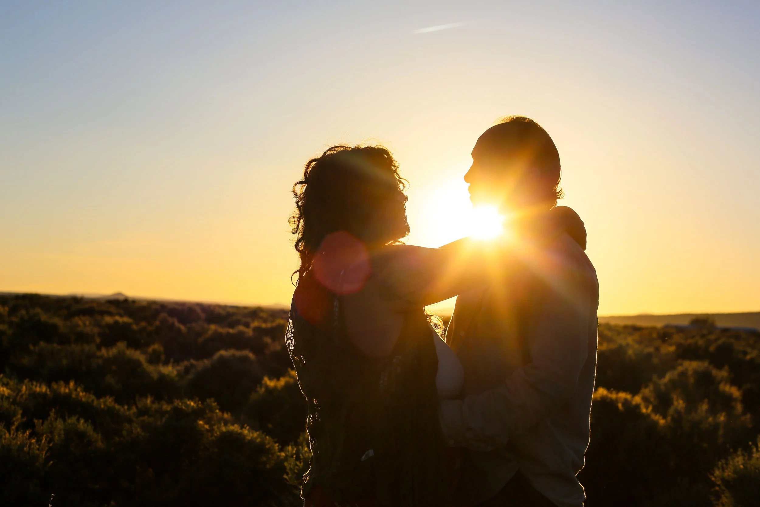 A couple embraces while facing each other at sunset in an outdoor landscape, with the sun shining behind them.