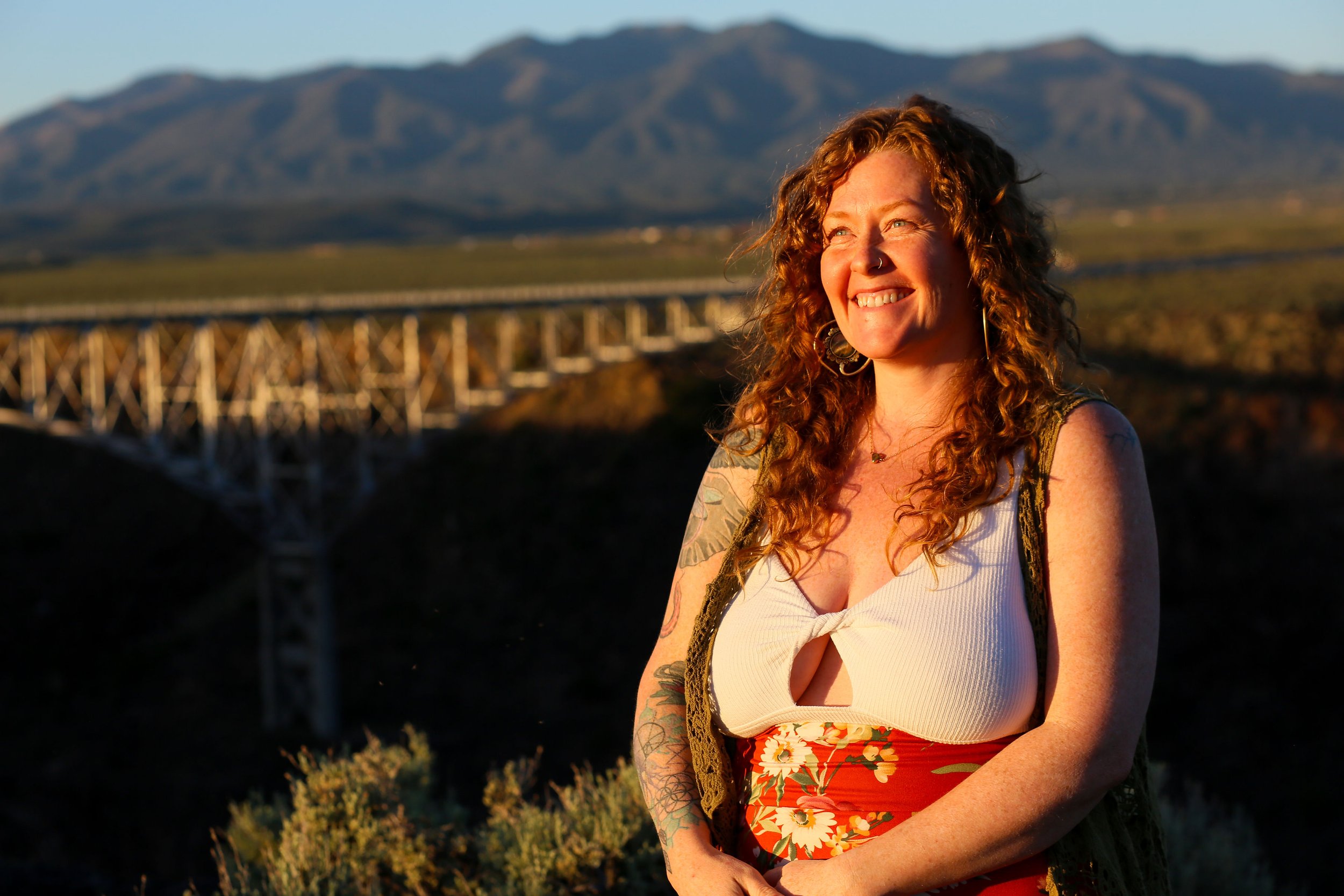A woman with curly red hair and tattoos on her arms, smiling and looking into the distance, standing outdoors during sunset with mountains and a bridge in the background.