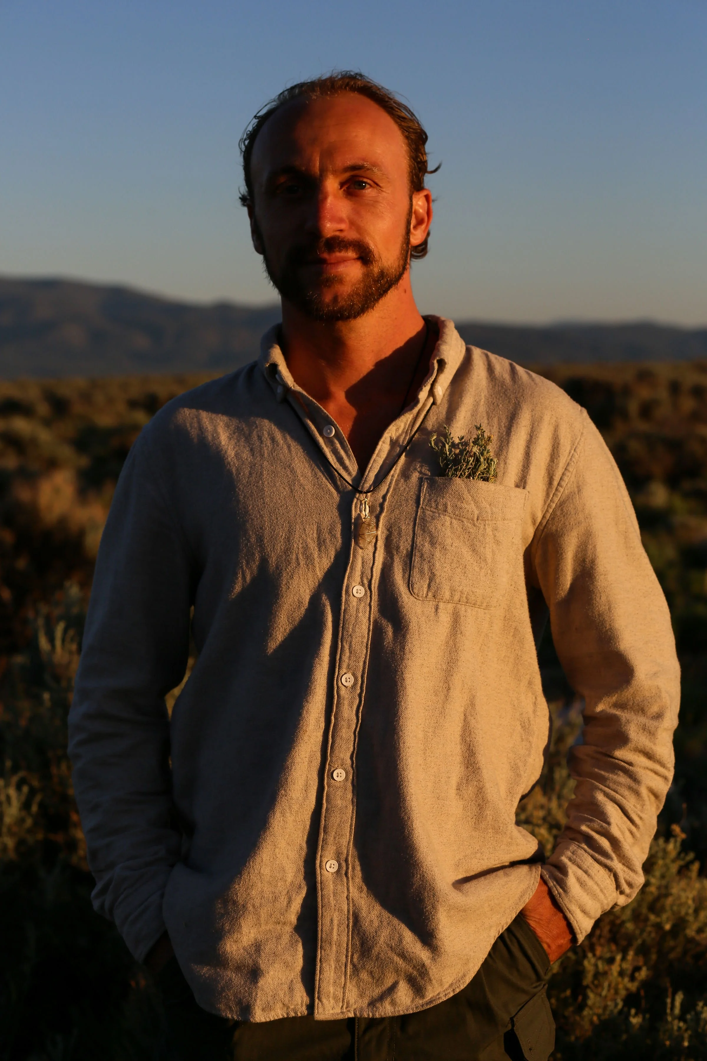A man with facial hair and a light-colored shirt standing outdoors during sunset, with mountains and desert landscape in the background.