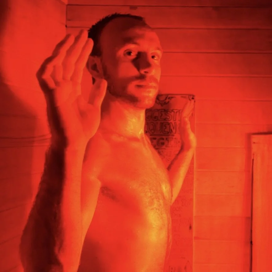 A shirtless man with short hair and a beard salutes, facing the camera, in a room with red lighting and wooden walls.