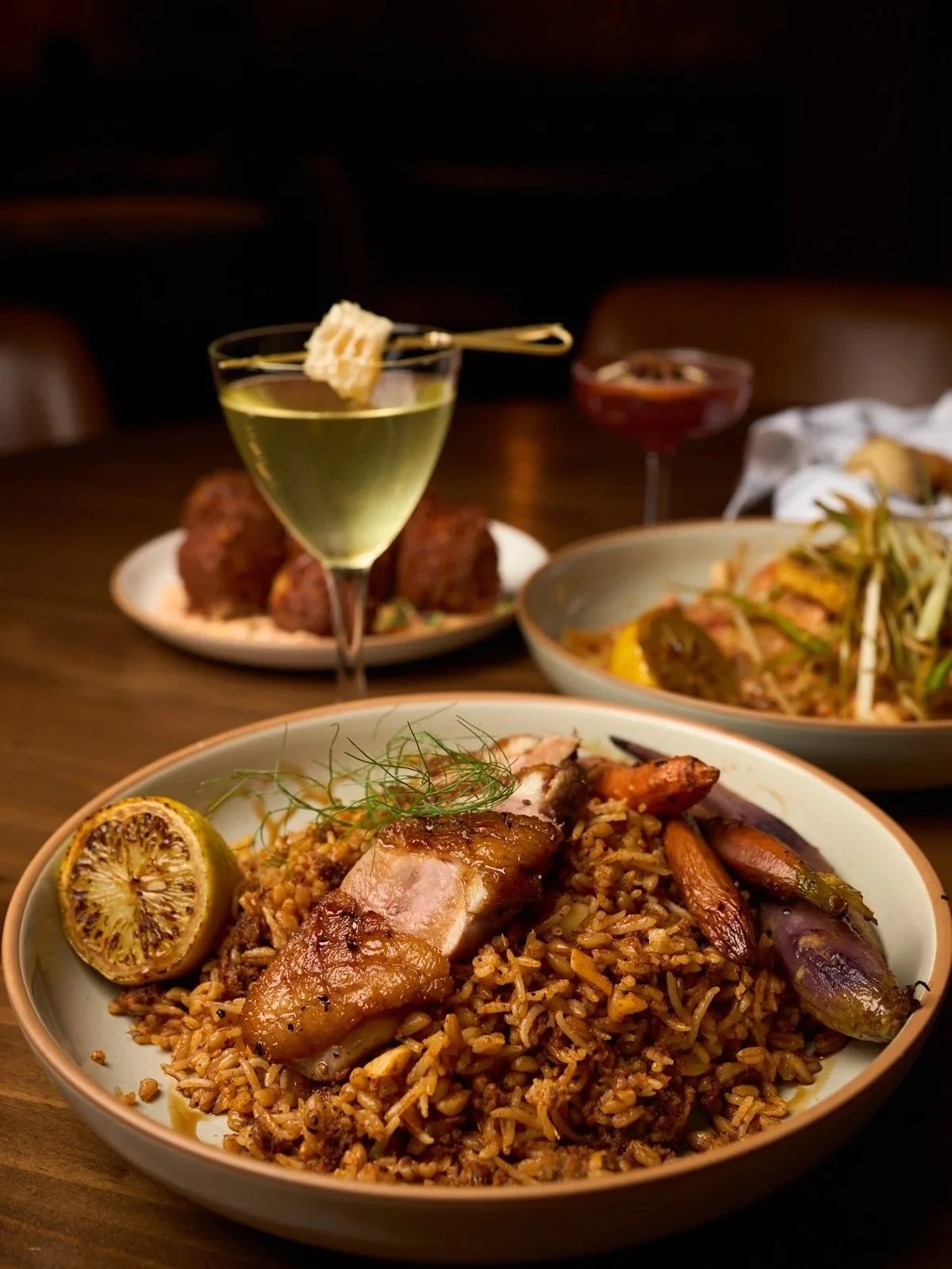 A main dish of fried rice topped with grilled chicken, garnished with a lime wedge and herbs, accompanied by various plates of food and drinks in the background.