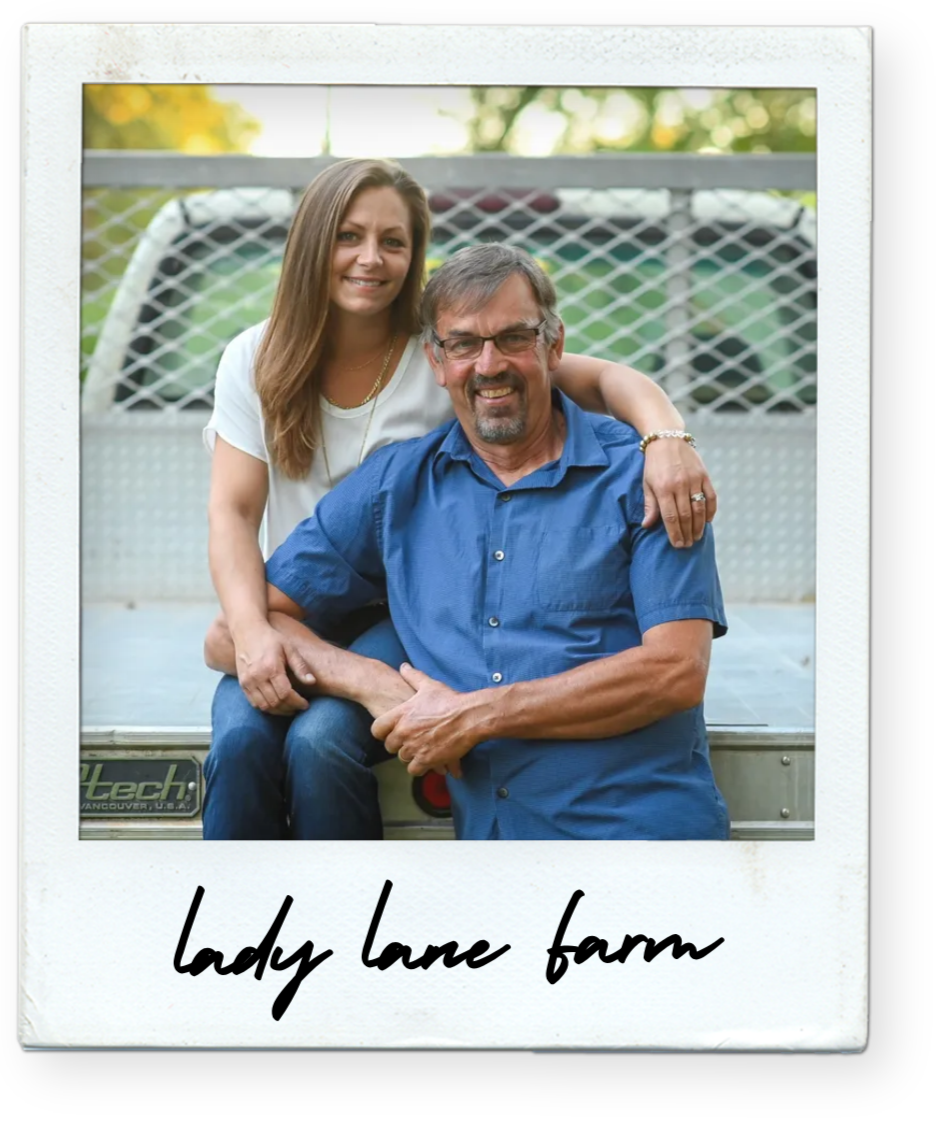 A woman with long brown hair and a man with glasses and a beard sitting on a trailer, smiling, with a fence and trees in the background, during daytime. The woman has her arm around the man, and they are outdoors at Lady Lane Farm.