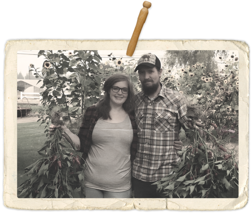 A black and white photo of a smiling woman (Ashley Hunt) and man (Joe Hunt) standing in a garden, holding large clusters of freshly picked vegetables, with sunflower plants in the background. The photo is styled like an old Polaroid picture.