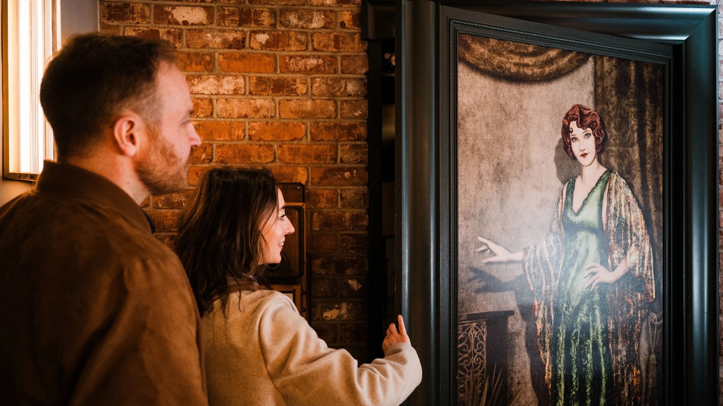 People peering behind a framed portrait of a woman with curly red hair in an art gallery with exposed brick walls. This is the secret entrance for the speakeasy below The Hive Social called The Hardware Store.