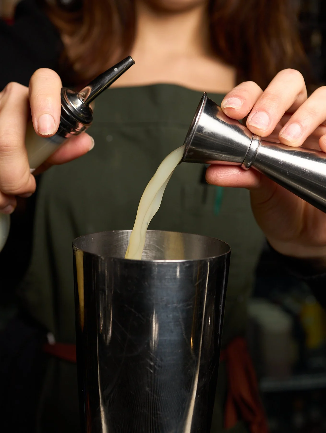 A person pouring a drink from a cocktail shaker into a metal glass, holding an open bottle in one hand and a shaker in the other.