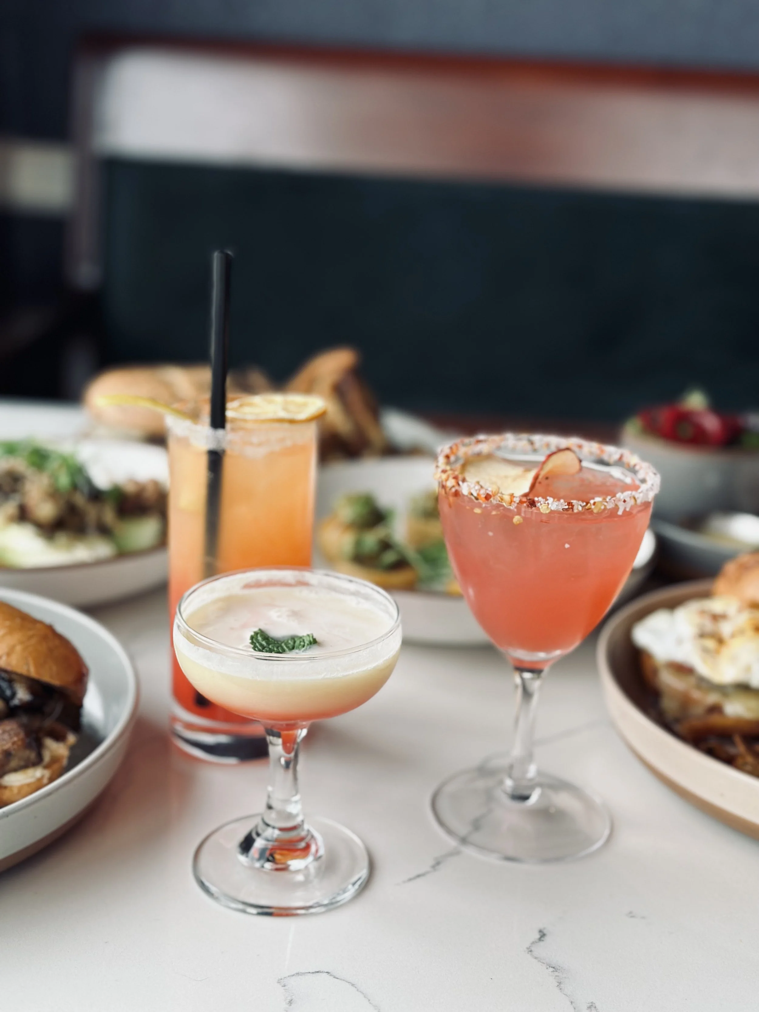 Close-up of three cocktails on a white table, with various dishes blurred in the background, including bowls of food and appetizers.