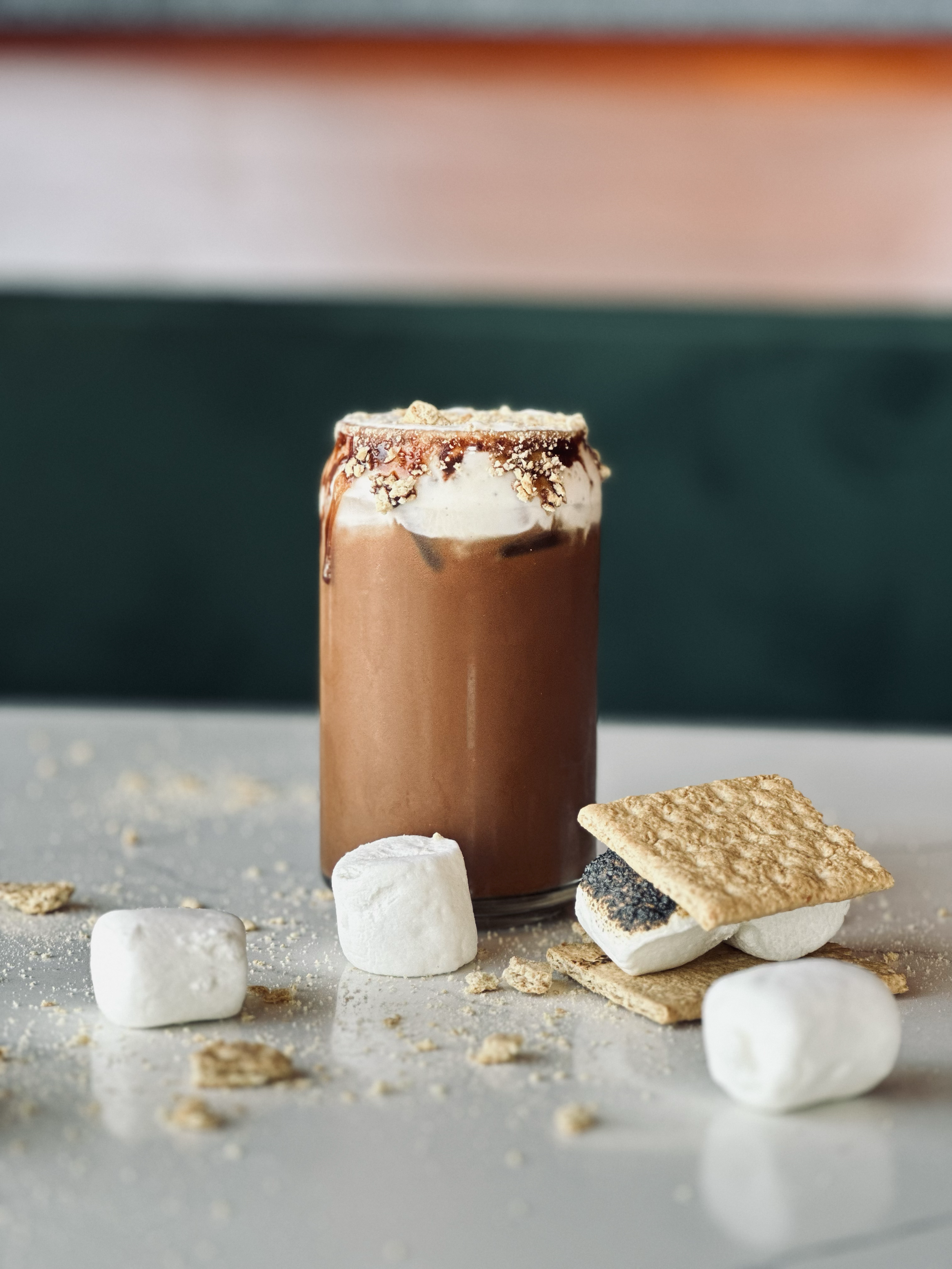 A smores cold brew, surrounded by marshmallows, graham crackers, and cookie pieces on a table.