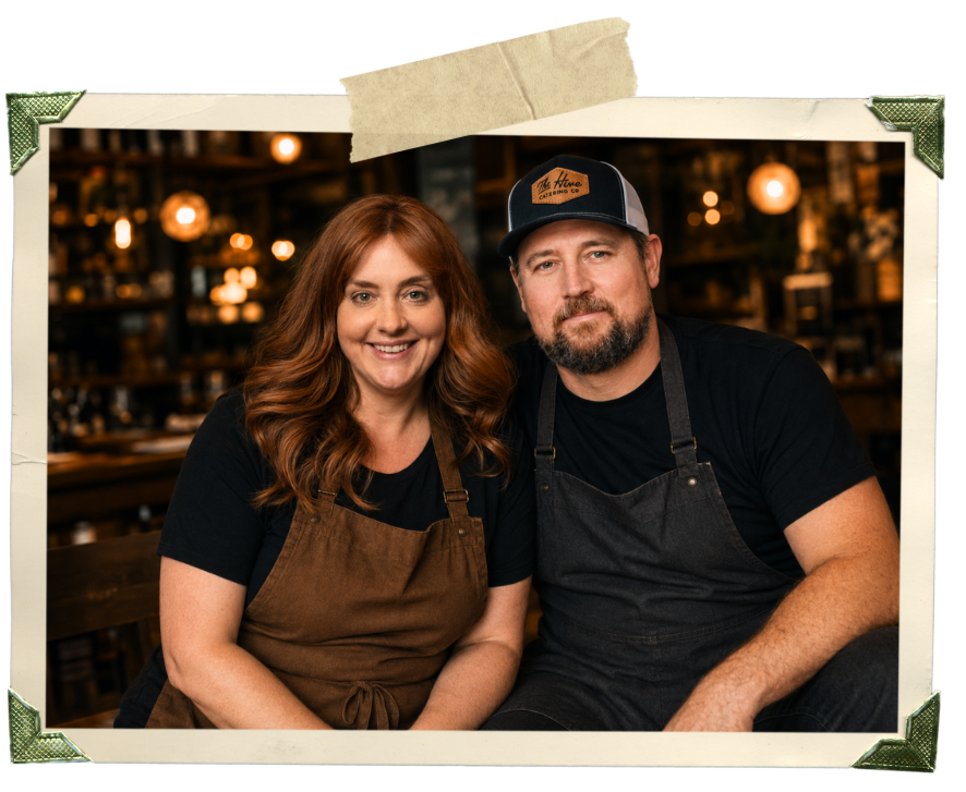 A smiling woman and a man sitting close together in a cozy restaurant or café. Both are wearing aprons, and the man has a trucker hat. The background has warm lighting and wooden shelves with various items.