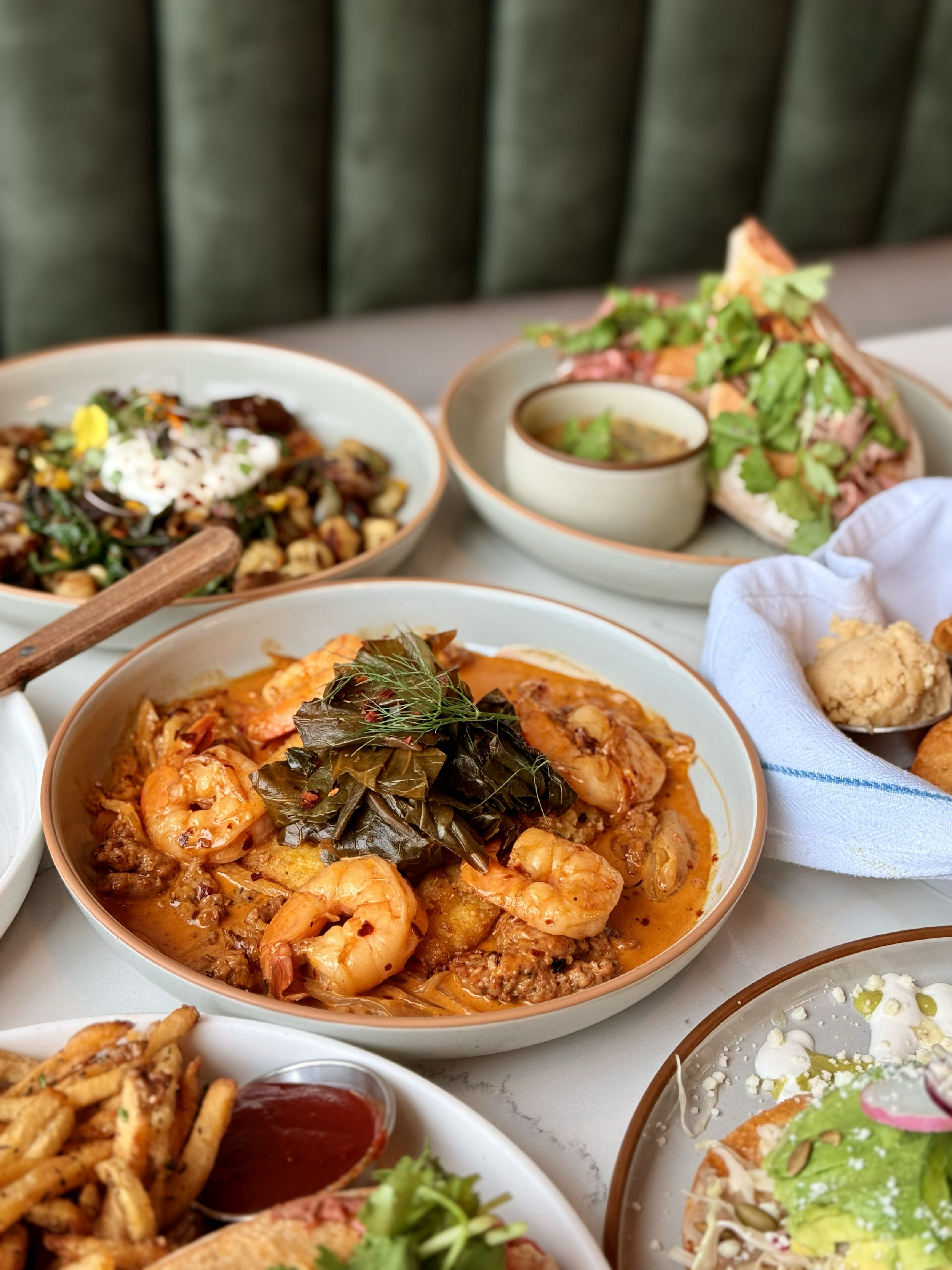 Various dishes including a shrimp and meat stew garnished with herbs, a salad with greens and herbs, a plate of pasta with herbs and tomato sauce, and a side of dipping sauce with ketchup.