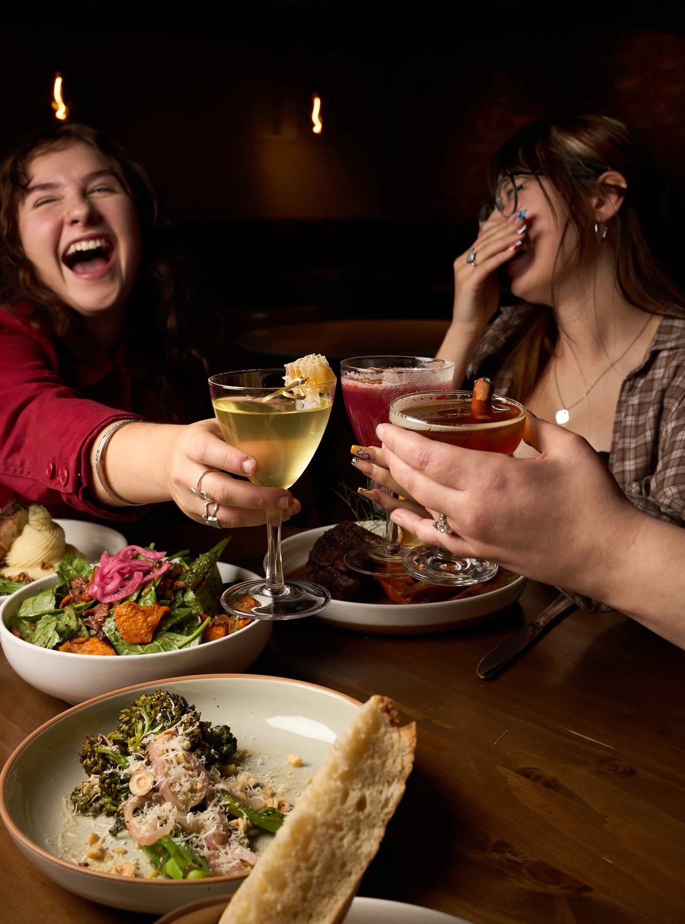 Two women smiling and laughing at a dinner table, holding cocktails in a dimly lit restaurant with food dishes including salad, pasta, and bread.