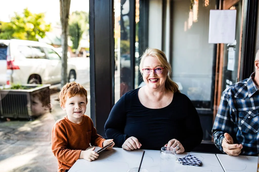 A smiling woman with glasses and red hair sitting at a table with two children, one boy wearing an orange sweater holding a phone, inside a restaurant or cafe with large glass windows showing parked cars outside.