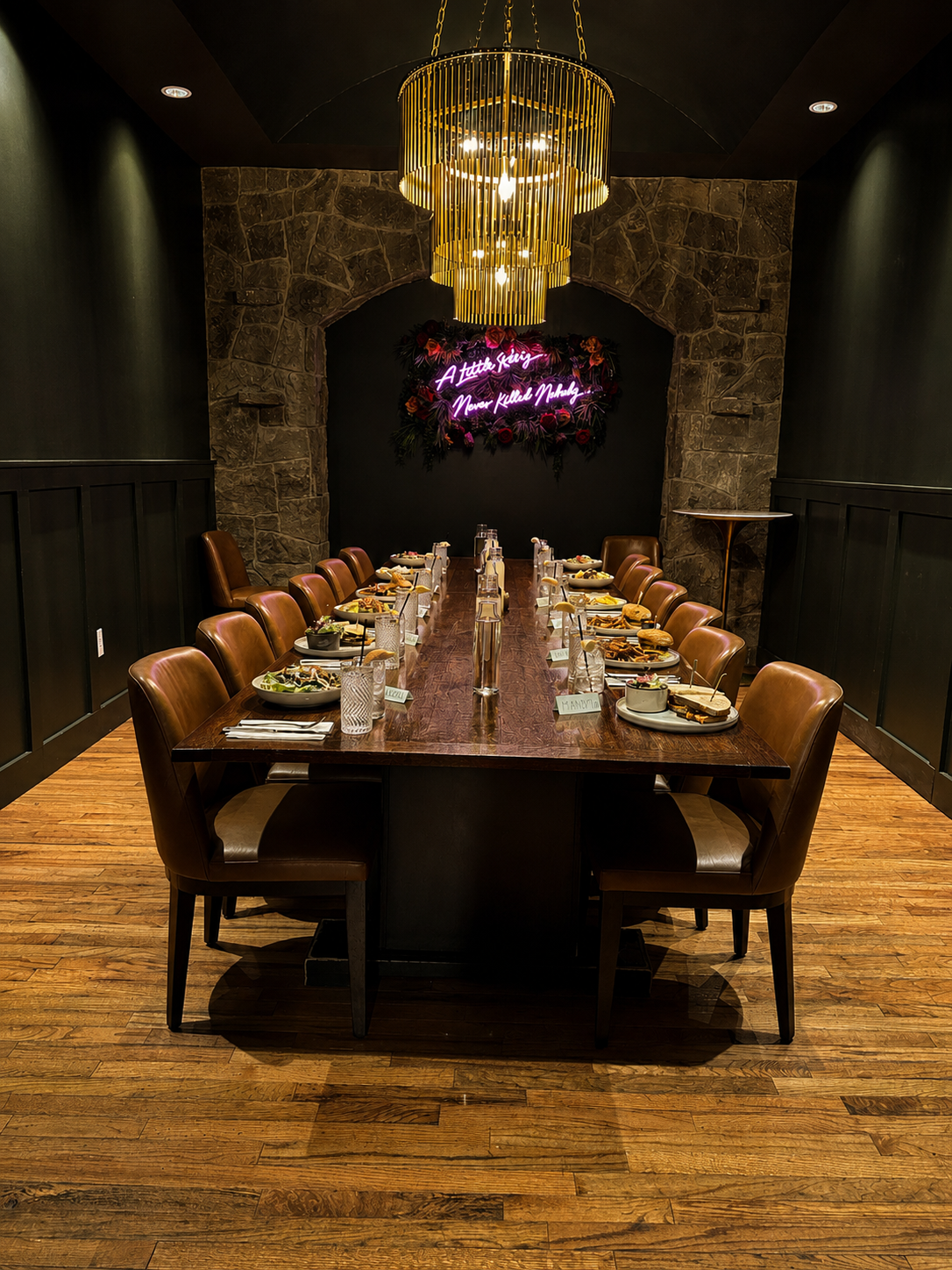 A long wooden dining table set for a meal in a dimly lit room with a dark stone brick wall. There are 12 chairs, six on each side, with plates of food, glasses, and utensils. This is the private dining room of the Hive Social.