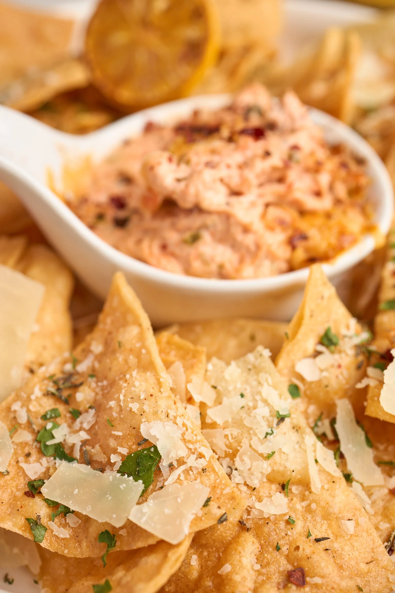 Close-up of nachos with shredded cheese, chopped herbs, and grated cheese on top, served with a side of spicy dip.