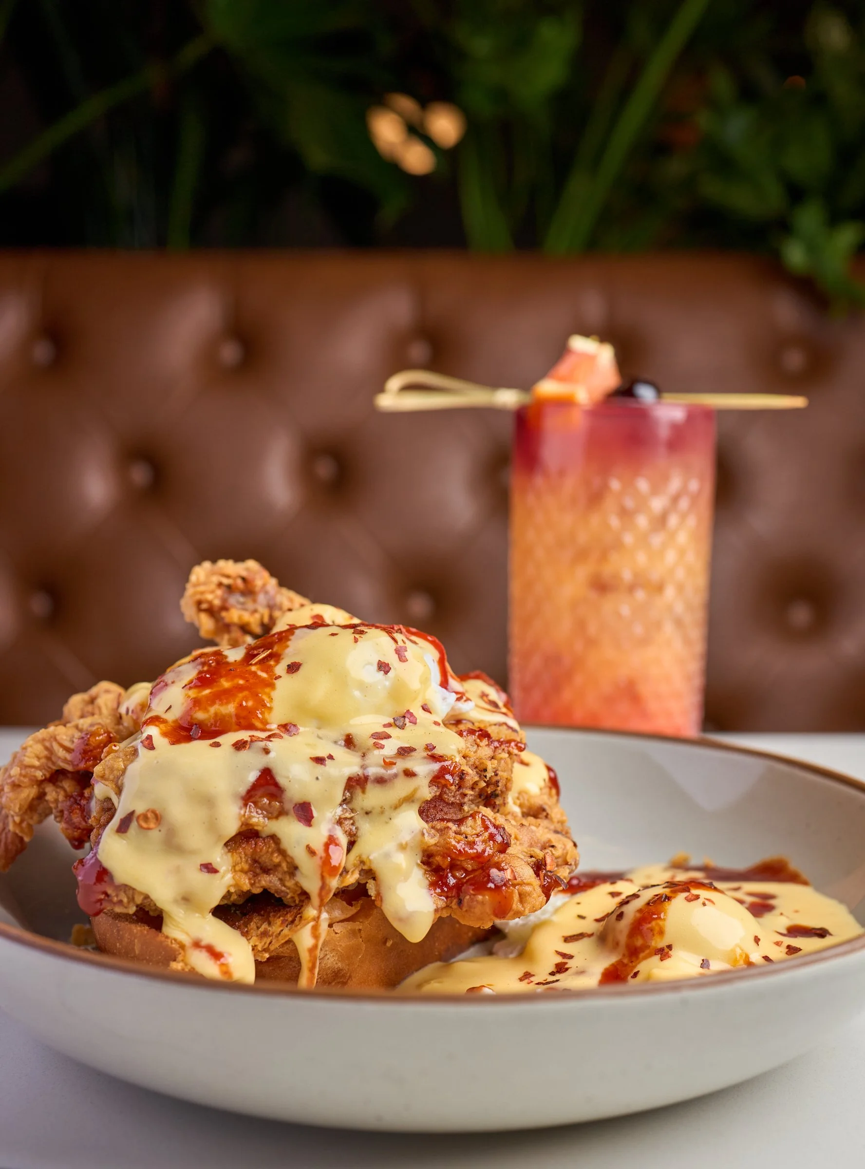 Close-up of a plate of fried chicken topped with melted cheese, red sauce, and red pepper flakes, with a blurred background featuring a tall drink with similar toppings and a decorative straw.