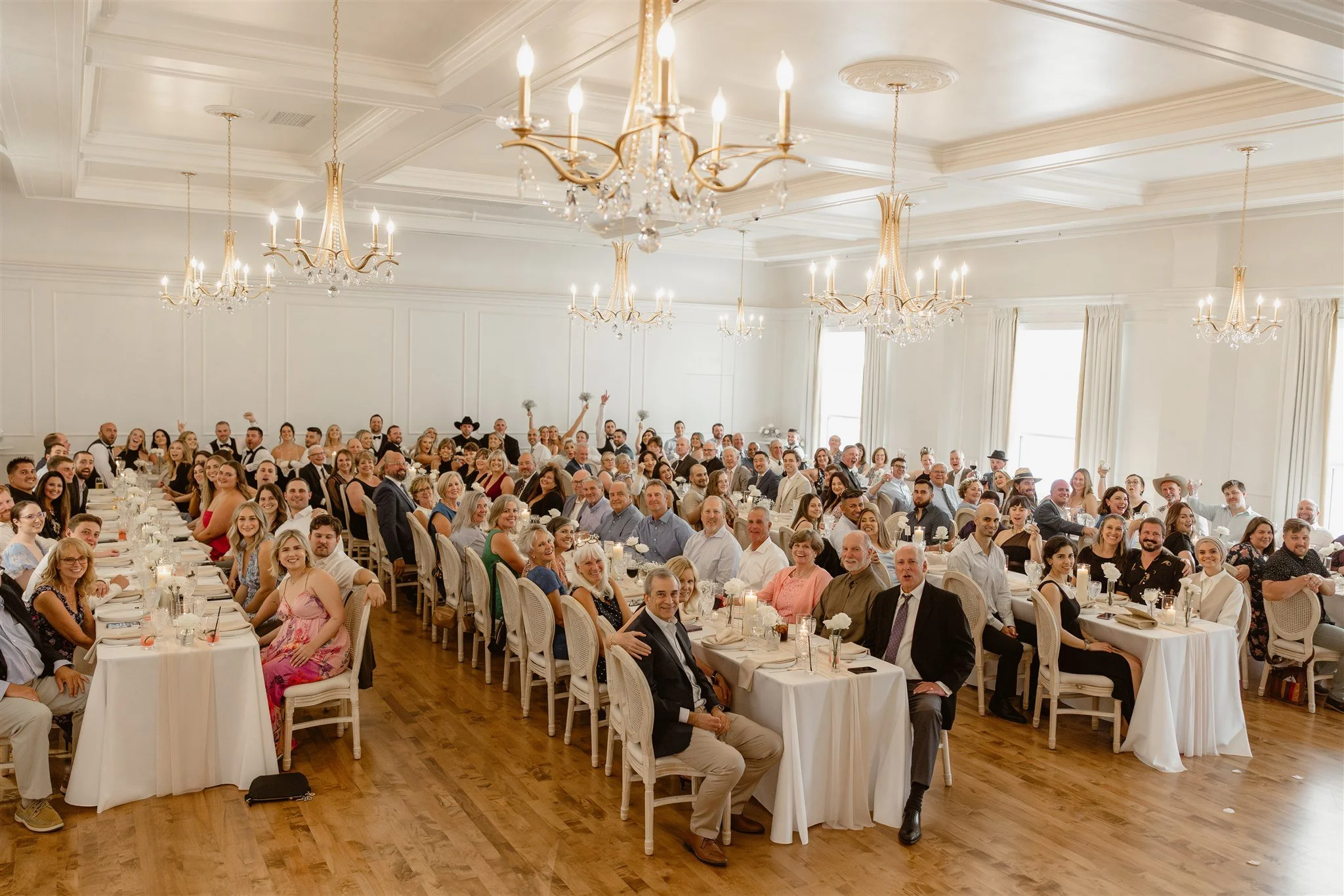 A large group of people gathered in a spacious, elegant banquet hall with high ceilings, large windows, and chandeliers. This is the grand ballroom of The Arc Light Venue above The Hive Social.