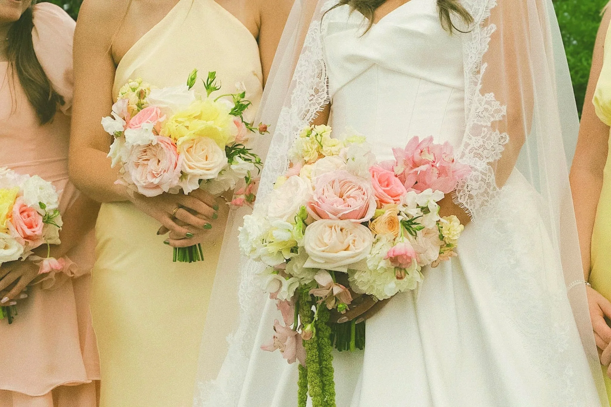 Close-up of a woman in a white dress holding a bouquet of pink, white, and yellow flowers with greenery, standing next to women in pastel-colored dresses holding similar bouquets, outdoors with greenery in the background.