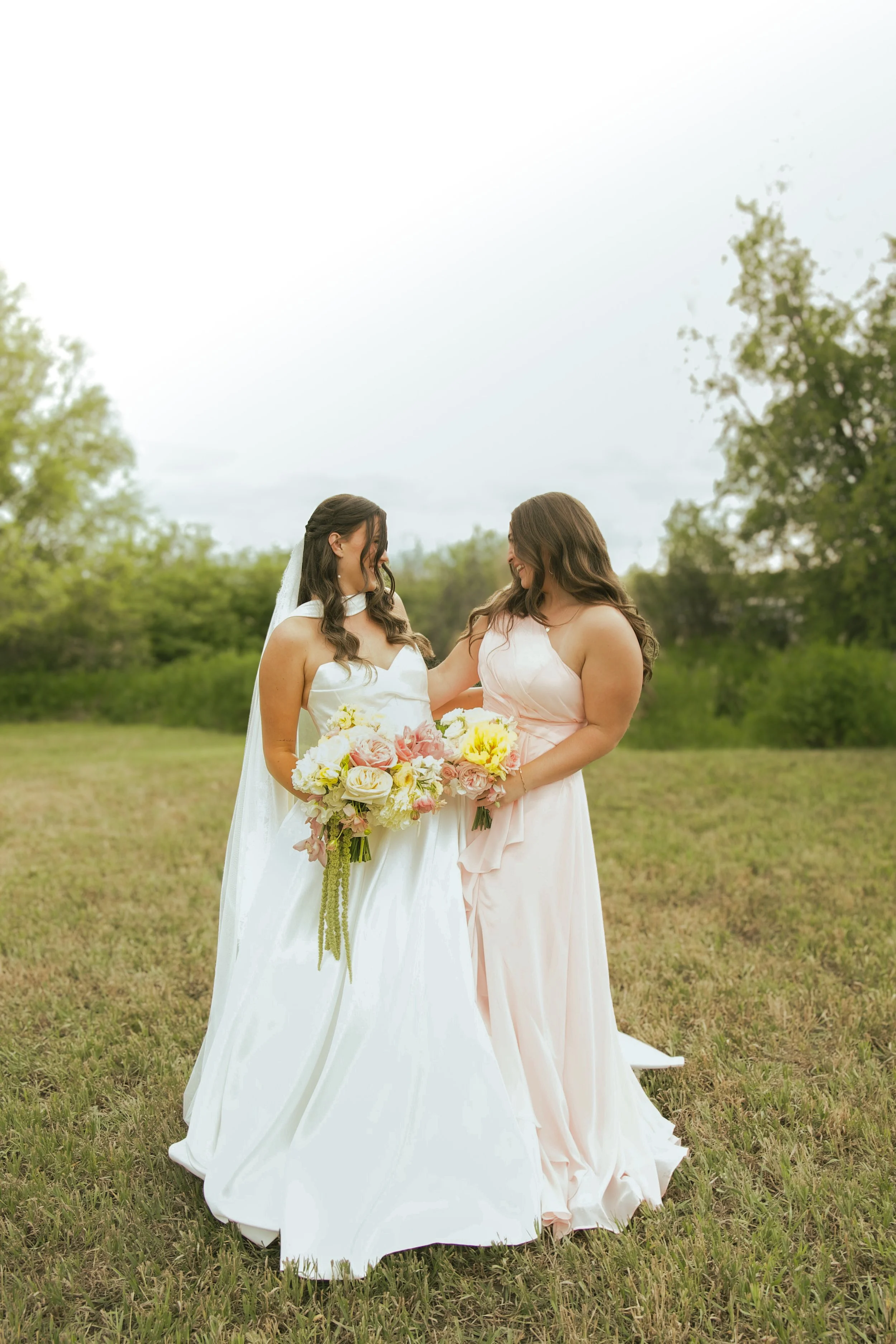 Two women in wedding dresses sharing a moment outdoors, one holding a large bouquet of flowers, surrounded by trees and grass.