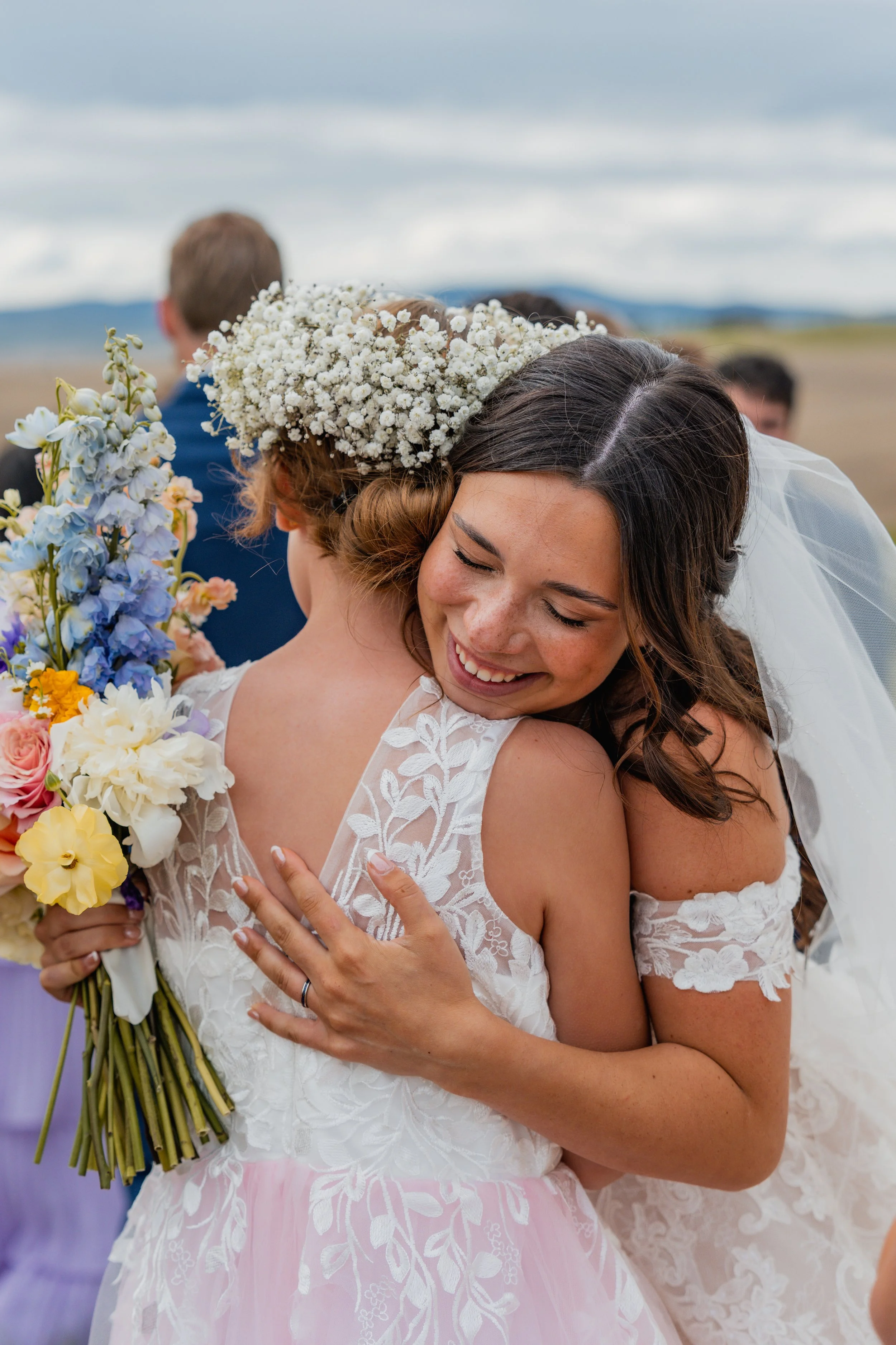 Two women sharing a warm hug at a wedding. One woman is holding a bouquet of flowers and is wearing a lace dress with pink accents. The other woman is wearing a lace dress with off-the-shoulder sleeves and a veil. People are in the background under a