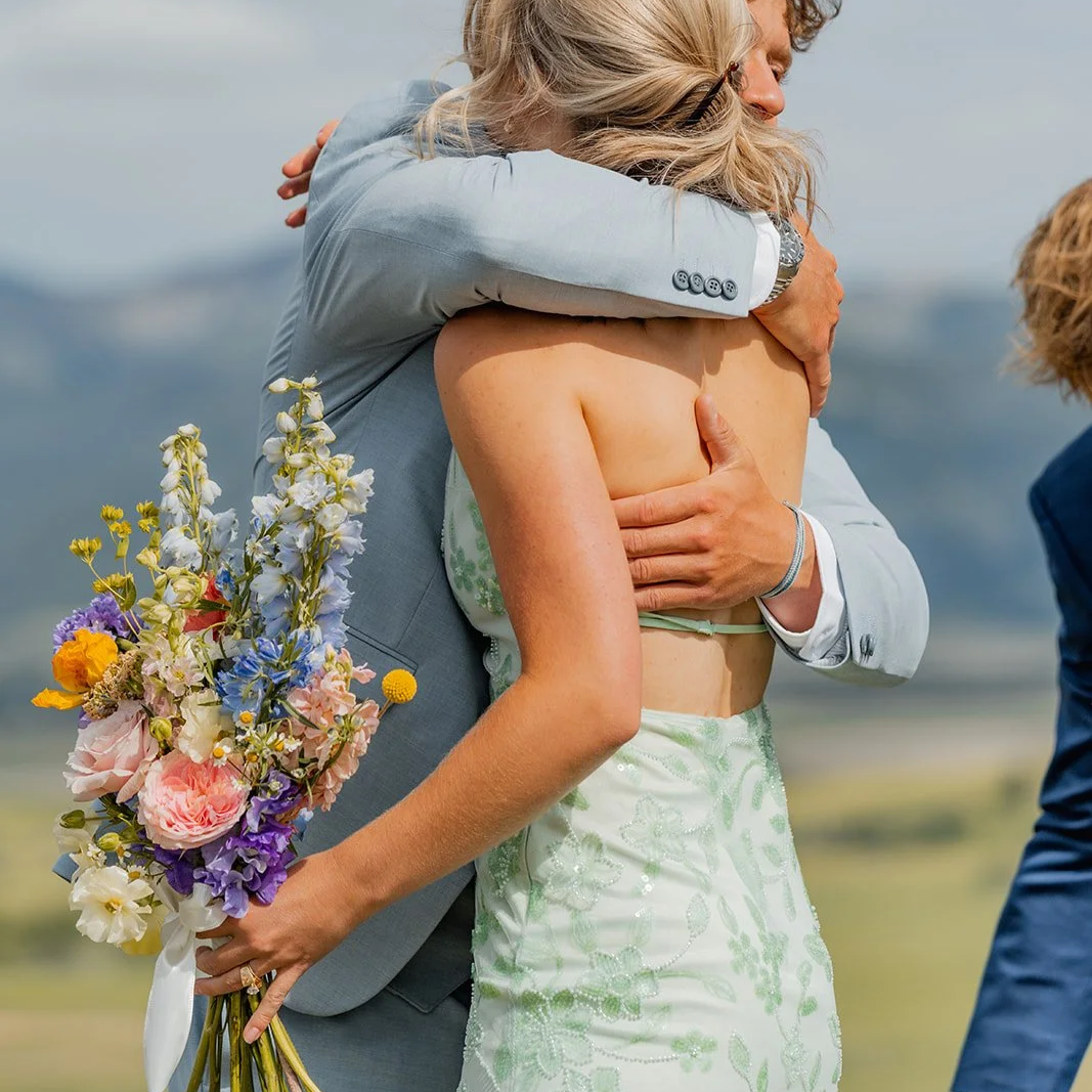 A couple embraces during their wedding ceremony, with the groom holding a bouquet of colorful flowers behind the bride. The background features a scenic outdoor landscape with mountains and a cloudy sky.