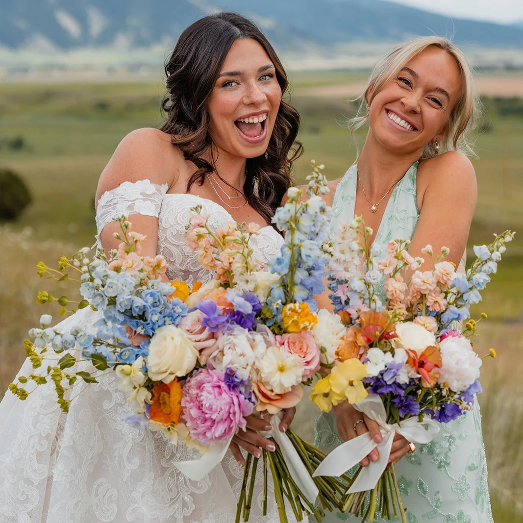 Two women smiling and holding vibrant bouquets of flowers outdoors, with a scenic landscape of fields and mountains in the background.
