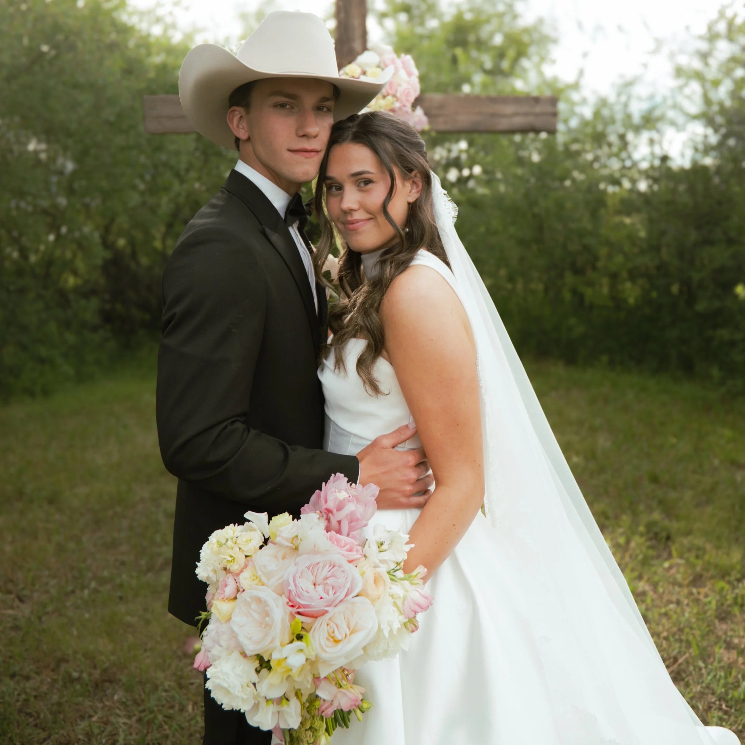 A young man in a tuxedo and large white cowboy hat and woman in a white dress, holding a bouquet of pink and white flowers, standing outdoors near a wooden cross with flowers, surrounded by greenery, during a wedding.