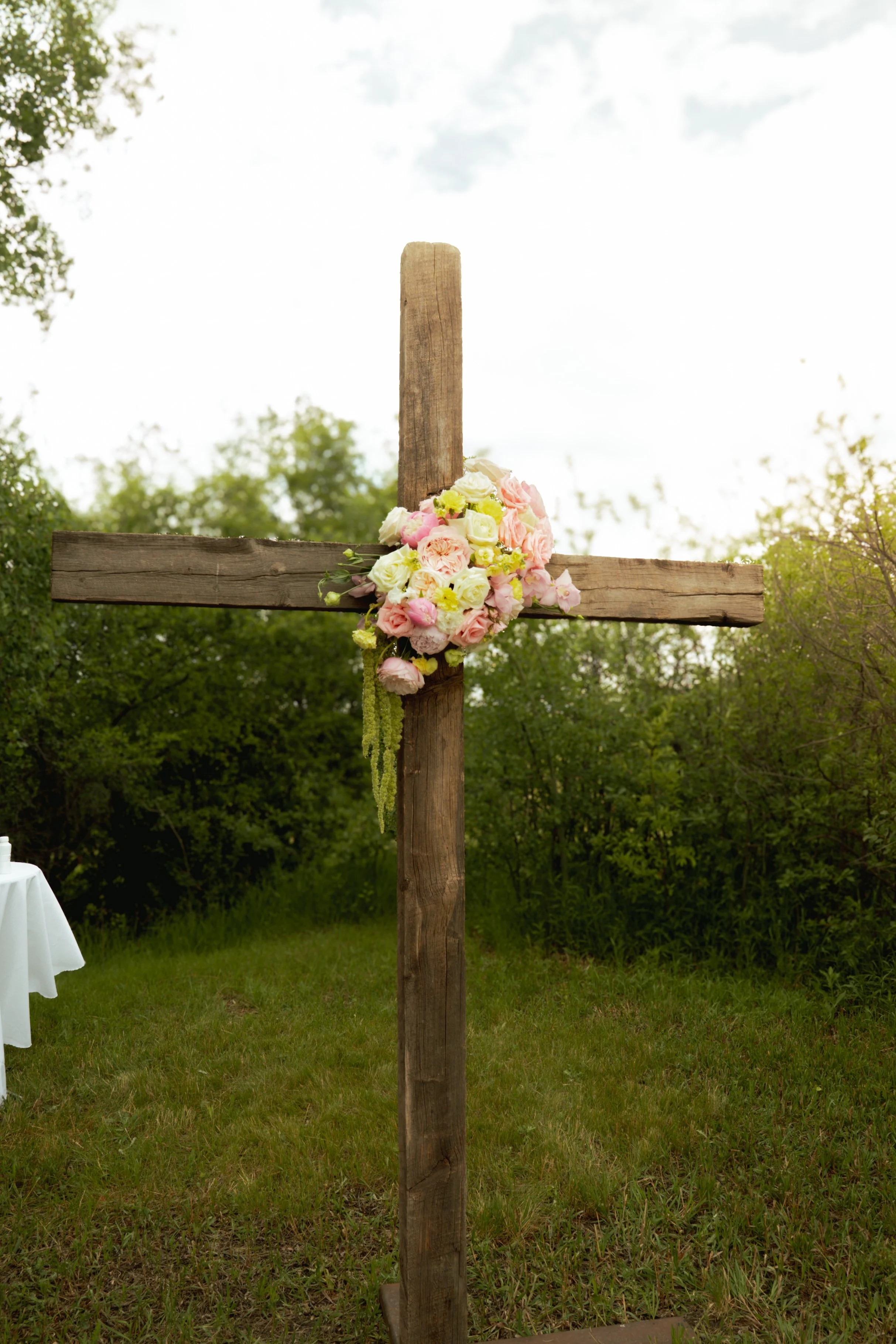Wooden cross decorated with pink and cream flowers, set outdoors with green trees and cloudy sky in the background.