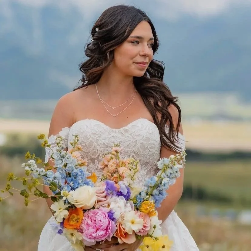 A bride in a white lace off-the-shoulder wedding dress holding a colorful bouquet of flowers outdoors with a scenic landscape background.