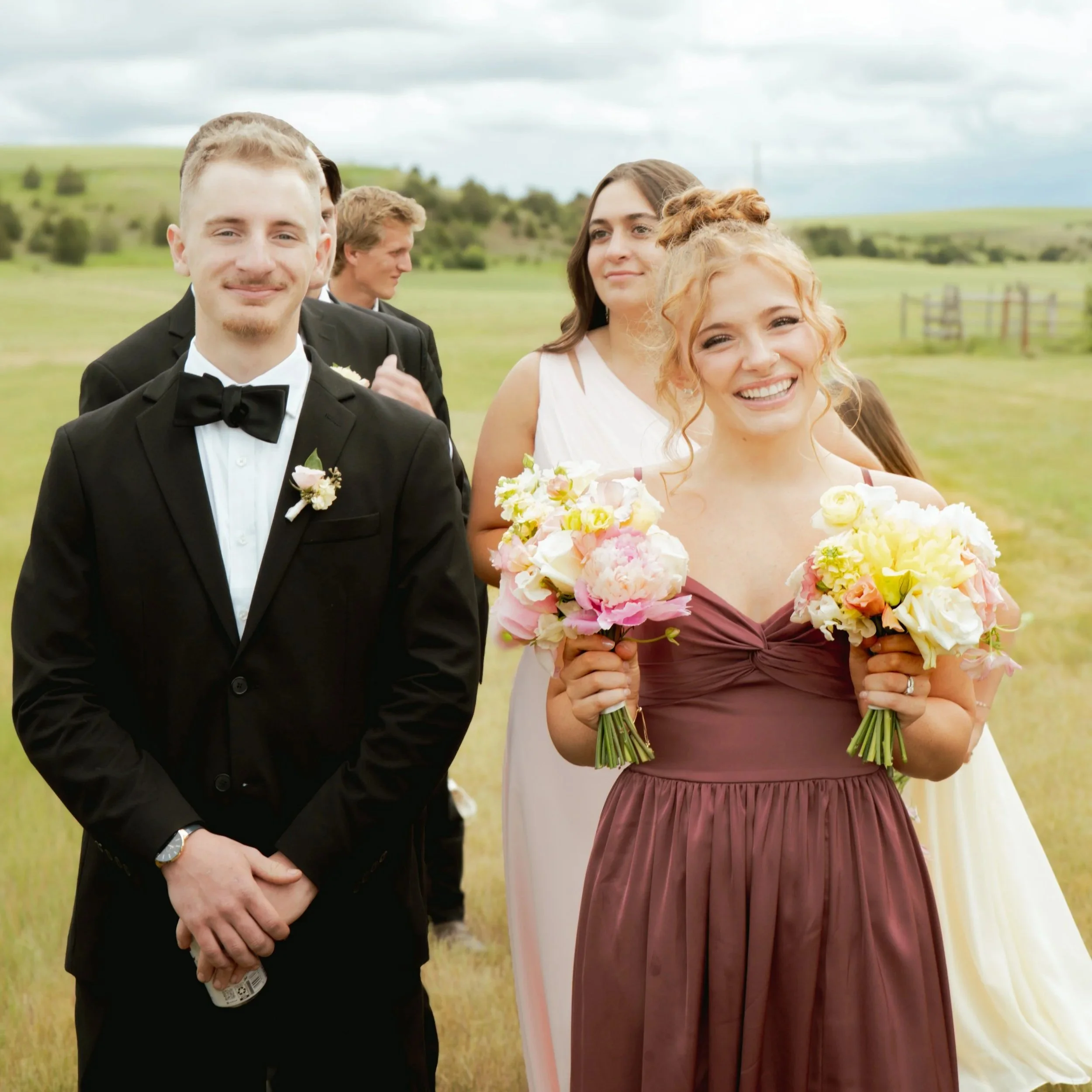 Group of people dressed in formal attire at an outdoor wedding ceremony in a grassy field, under cloudy skies.