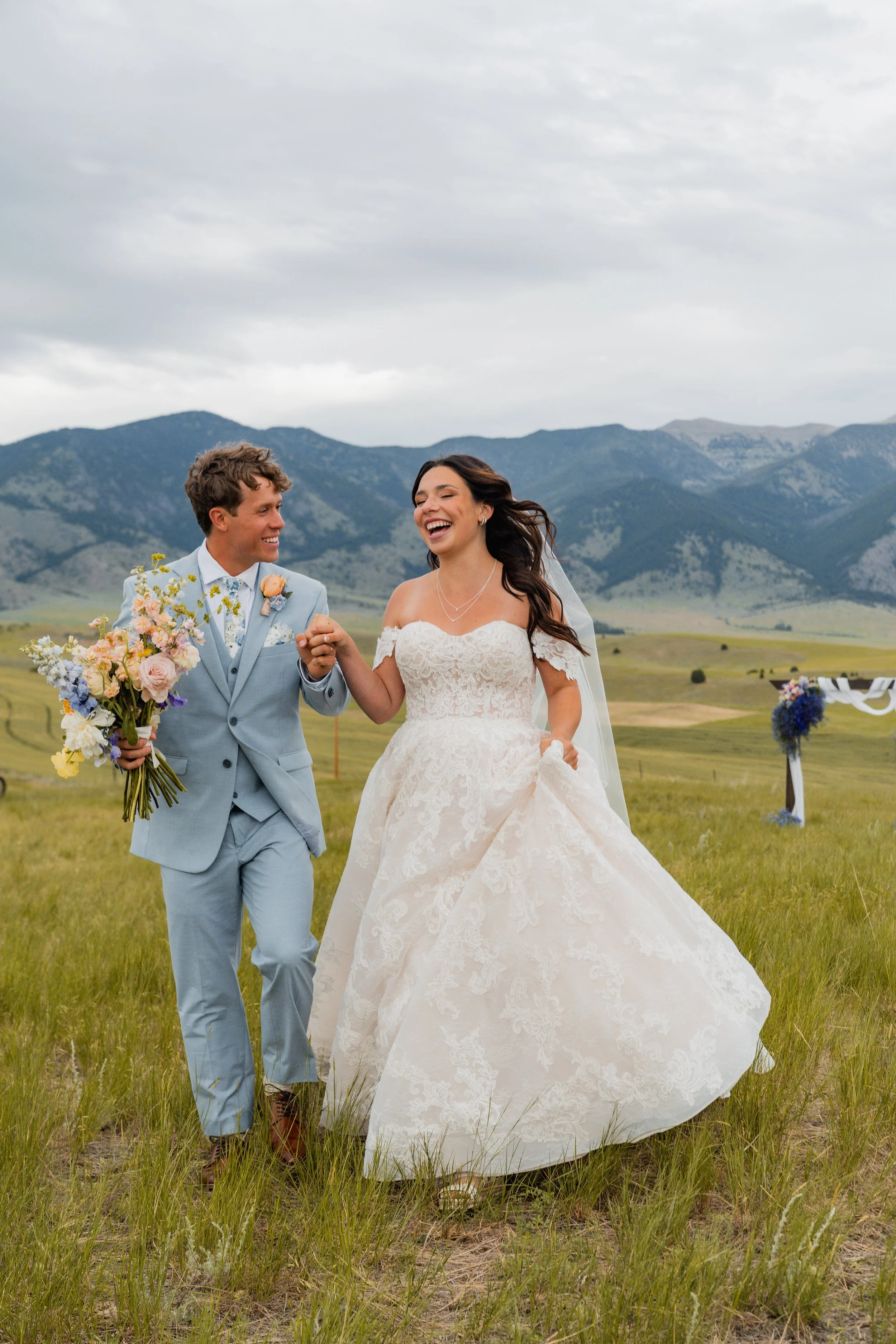 A joyful bride and groom dance outdoors in a grassy field with mountains in the background. The bride wears a white lace wedding dress, and the groom a light blue suit, holding a bouquet of flowers. They are smiling and holding hands.