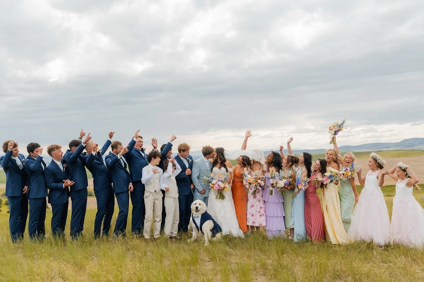 A wedding party standing in a grassy field with cloudy sky, including bridesmaids in colorful dresses, groomsmen in blue suits, a bride and groom, two flower girls, and a dog dressed in a suit, all celebrating and smiling.