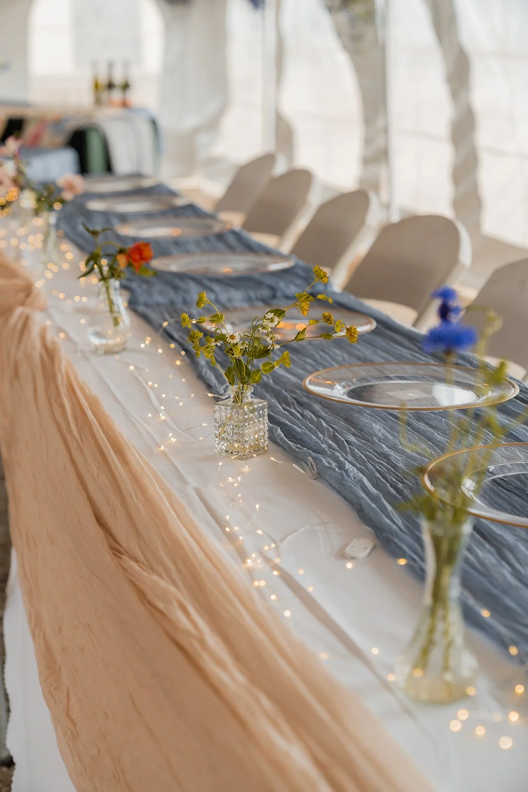 A long banquet table decorated with vases of flowers, a dark blue table runner, and lights, set for a meal in a bright room with large windows.
