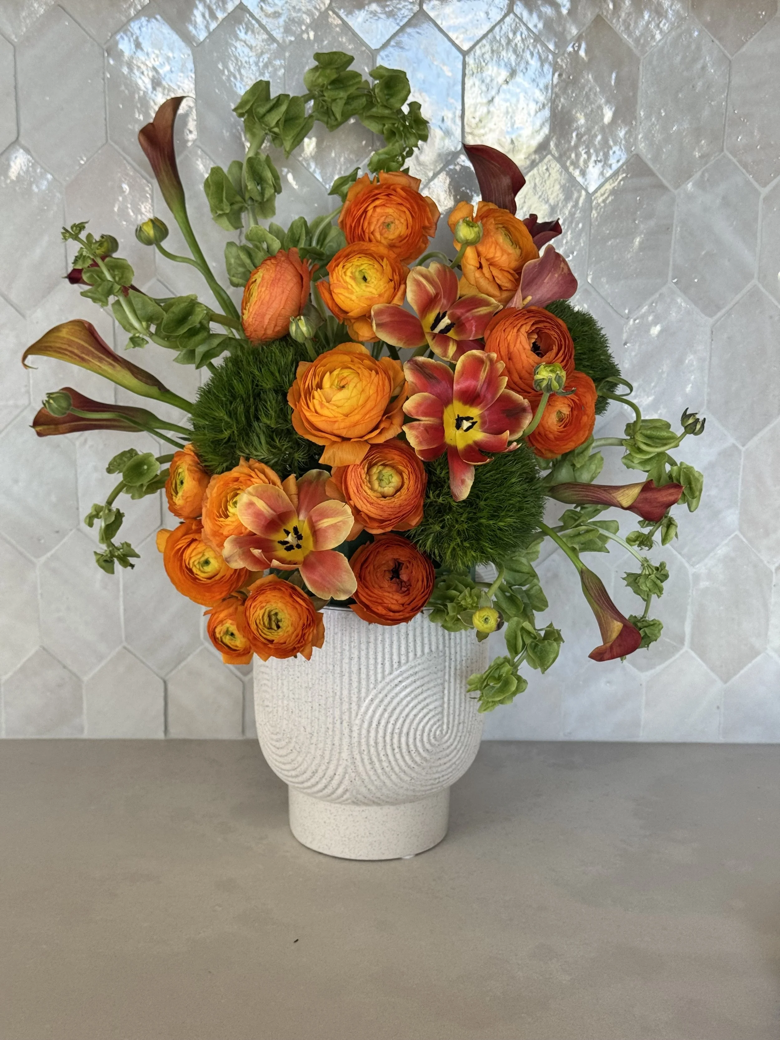 A white textured ceramic vase with a rounded base and narrow neck holding a colorful bouquet of flowers, placed on a light-colored flat surface in front of a white tiled wall with a hexagonal pattern.
