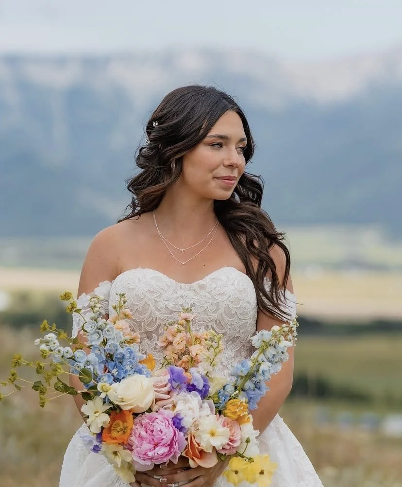 A bride holding a colorful bouquet of flowers outdoors, with a mountainous landscape in the background.