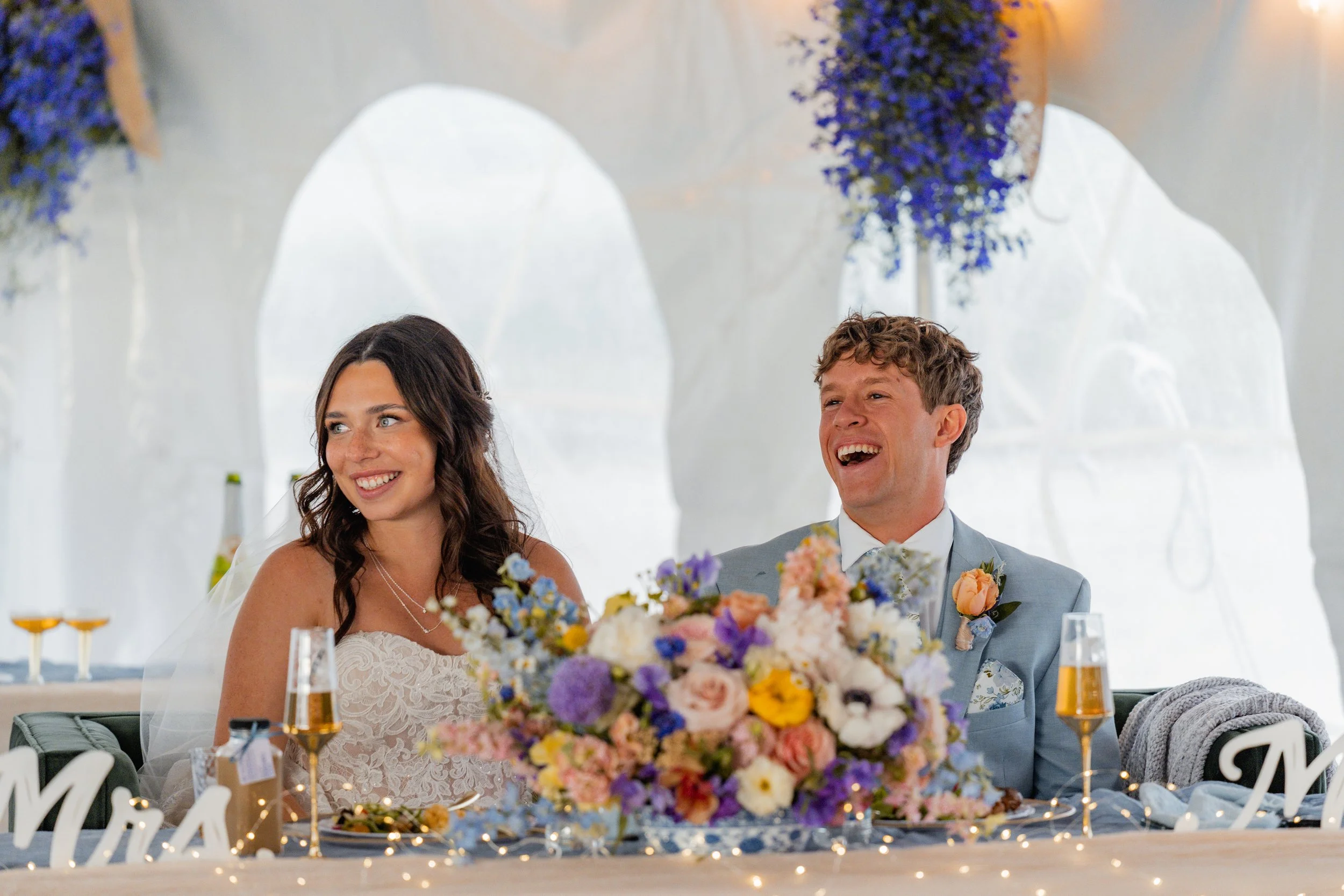 Smiling bride and groom sitting at a wedding reception table decorated with colorful flowers and surrounded by wedding guests.