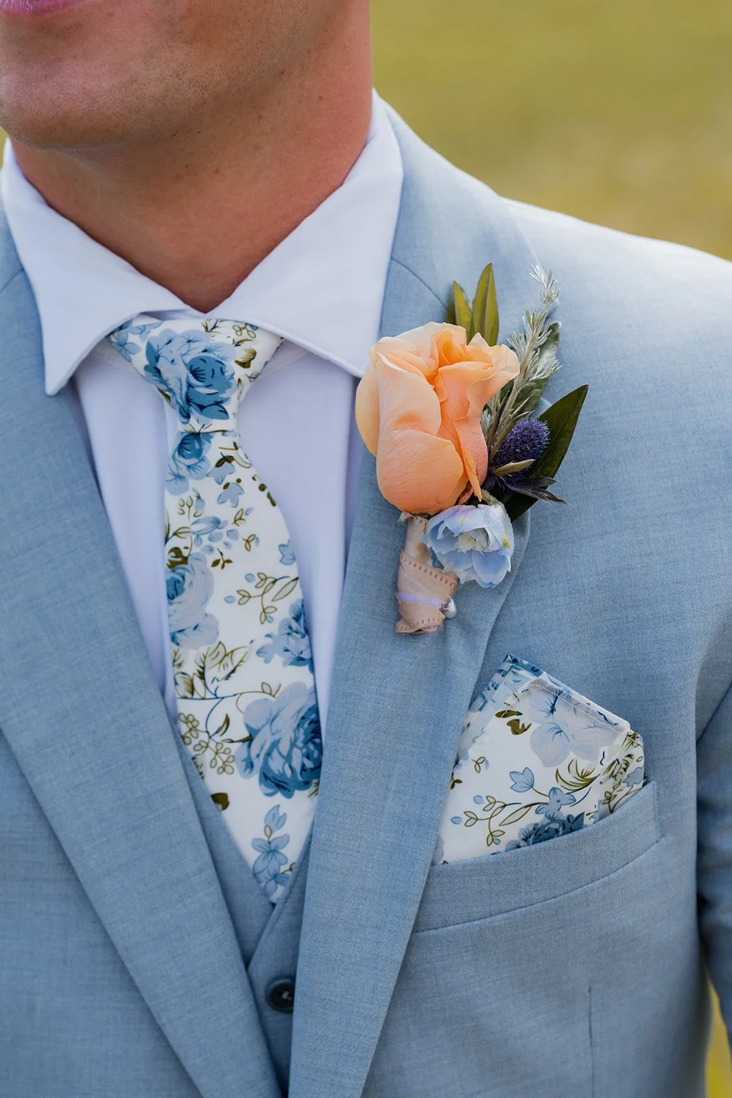 Close-up of a groom wearing a light grey suit with a floral tie, floral pocket square, and a boutonniere with peach and purple flowers on his lapel.