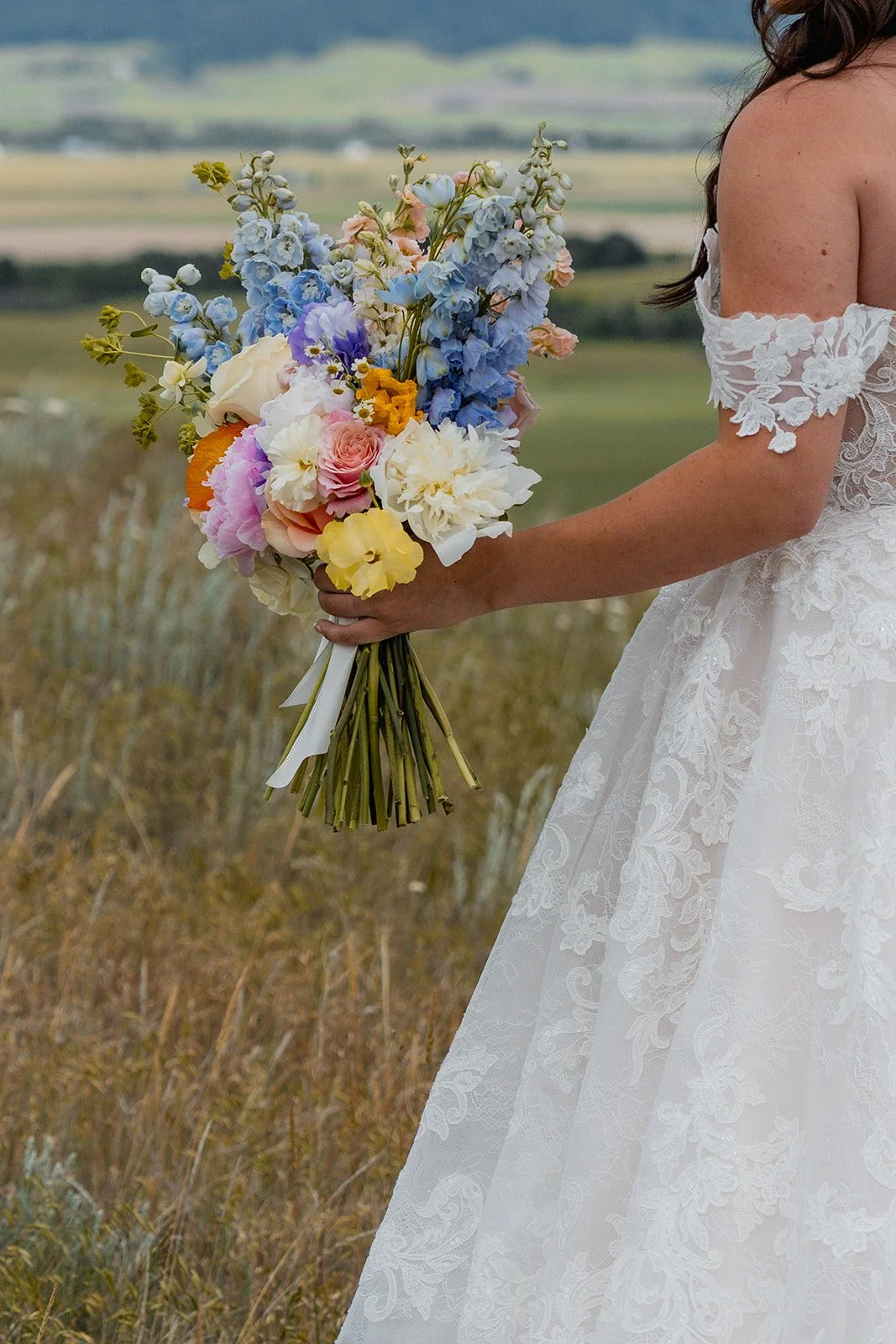 A woman in a lace wedding dress holding a colorful bouquet of flowers outdoors on a grassy field with a scenic landscape in the background.
