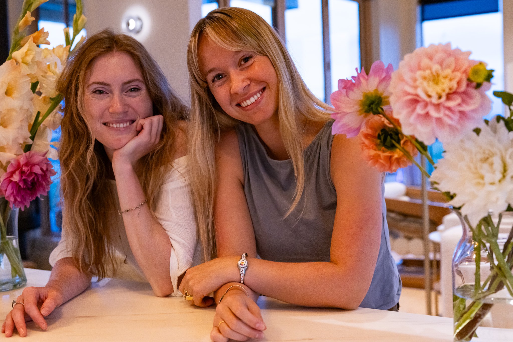 Two women smiling at the camera, sitting at a table surrounded by pink and cream flowers in vases, in a well-lit indoor space.