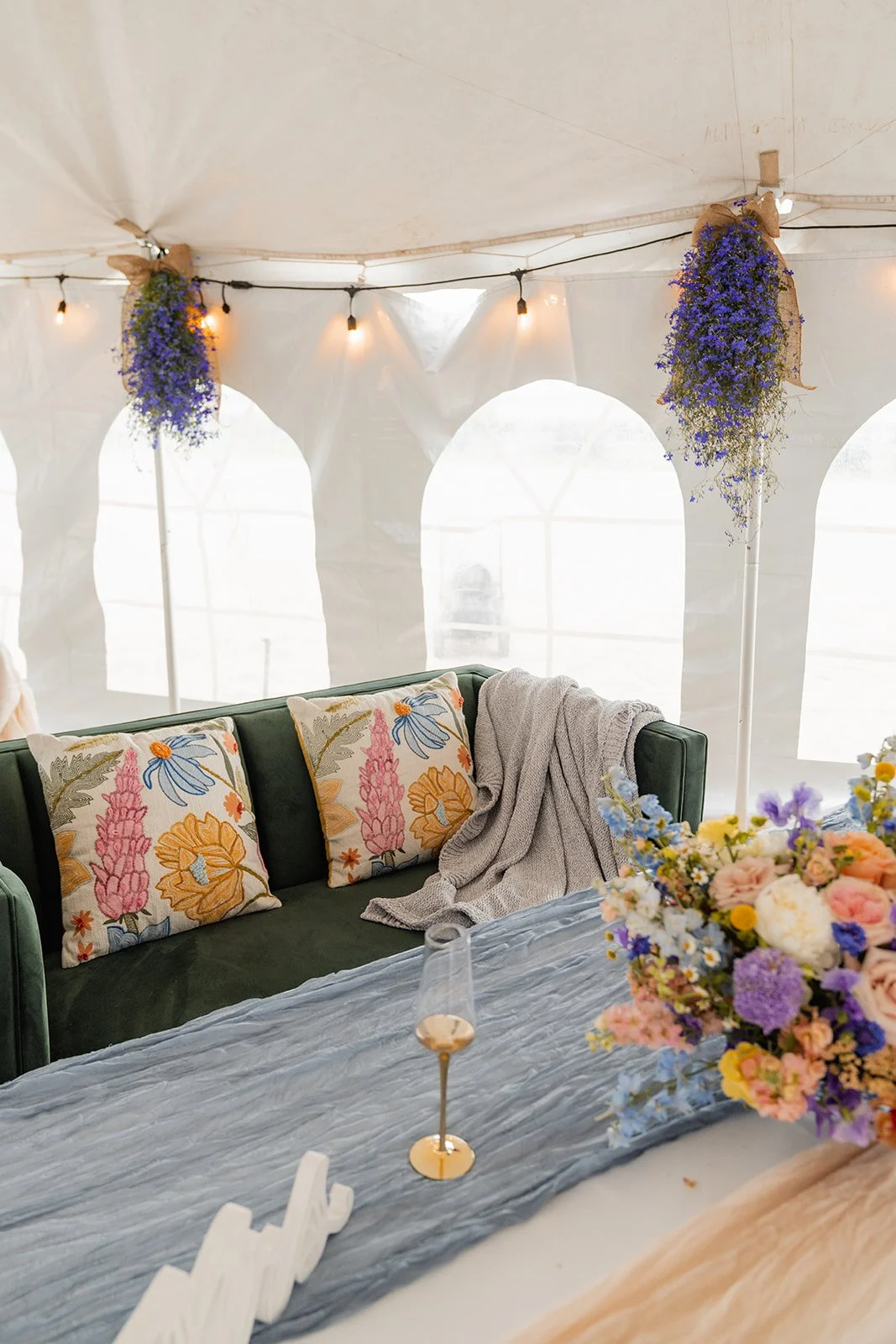 Interior of a decorated tent with string lights, floral arrangements, decorative pillows on a green couch, and a glass of wine on a table.