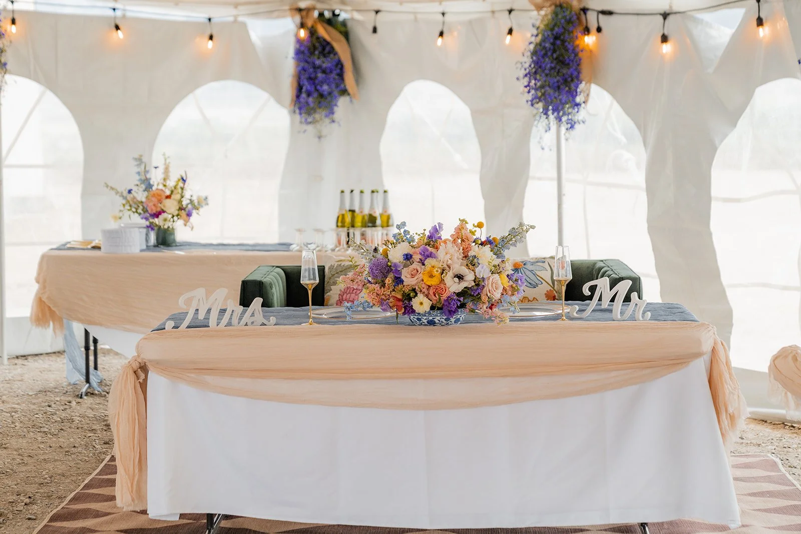 Decorated wedding reception table with a floral centerpiece, champagne glasses, and 'Mr.' and 'Mrs.' signs inside a tent with hanging string lights and blue floral arrangements.
