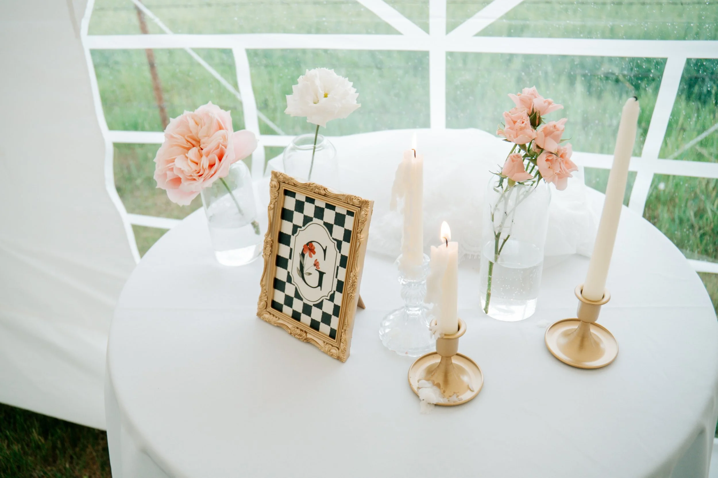 A round white table decorated with glass vases holding pink and white flowers, several white candles, a gold ornate picture frame with a black-and-white checkerboard pattern and the letter 'G', and cream-colored candleholders, set in front of a white