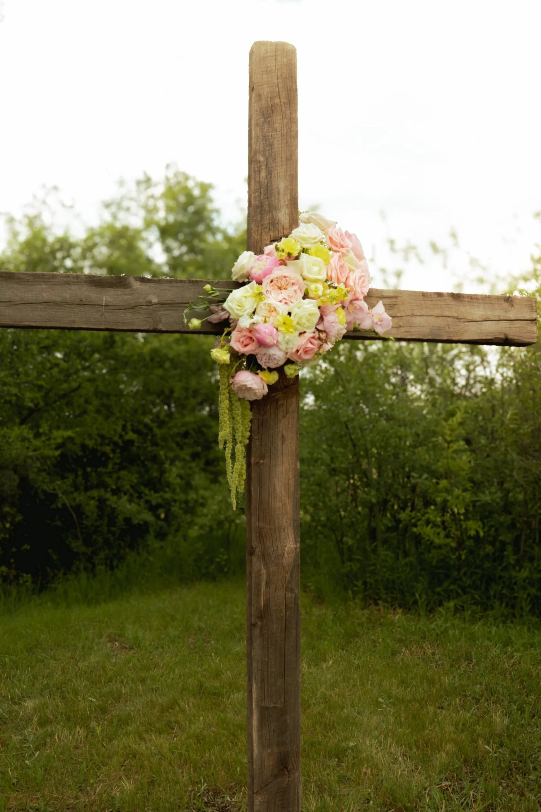 A wooden cross with a bouquet of pink, white, and yellow flowers hanging on it, outdoors with green trees and grass in the background.