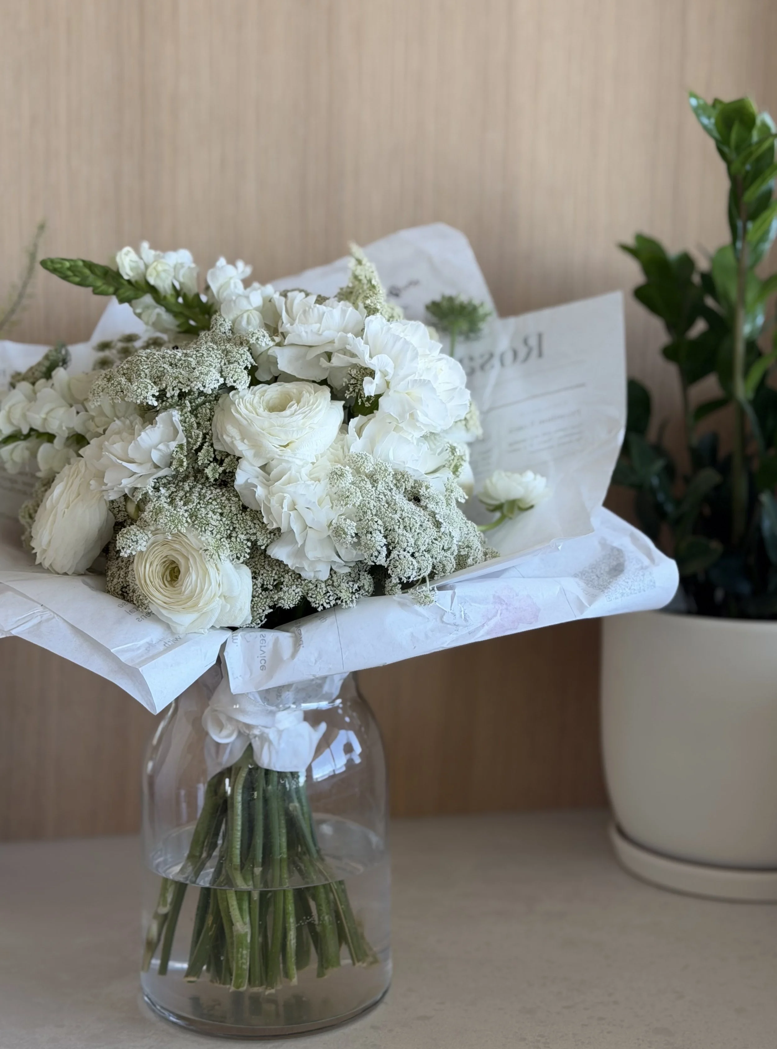 A bouquet of white flowers, including roses and greenery, arranged in a clear glass vase with water, placed on a light-colored surface against a wooden background.