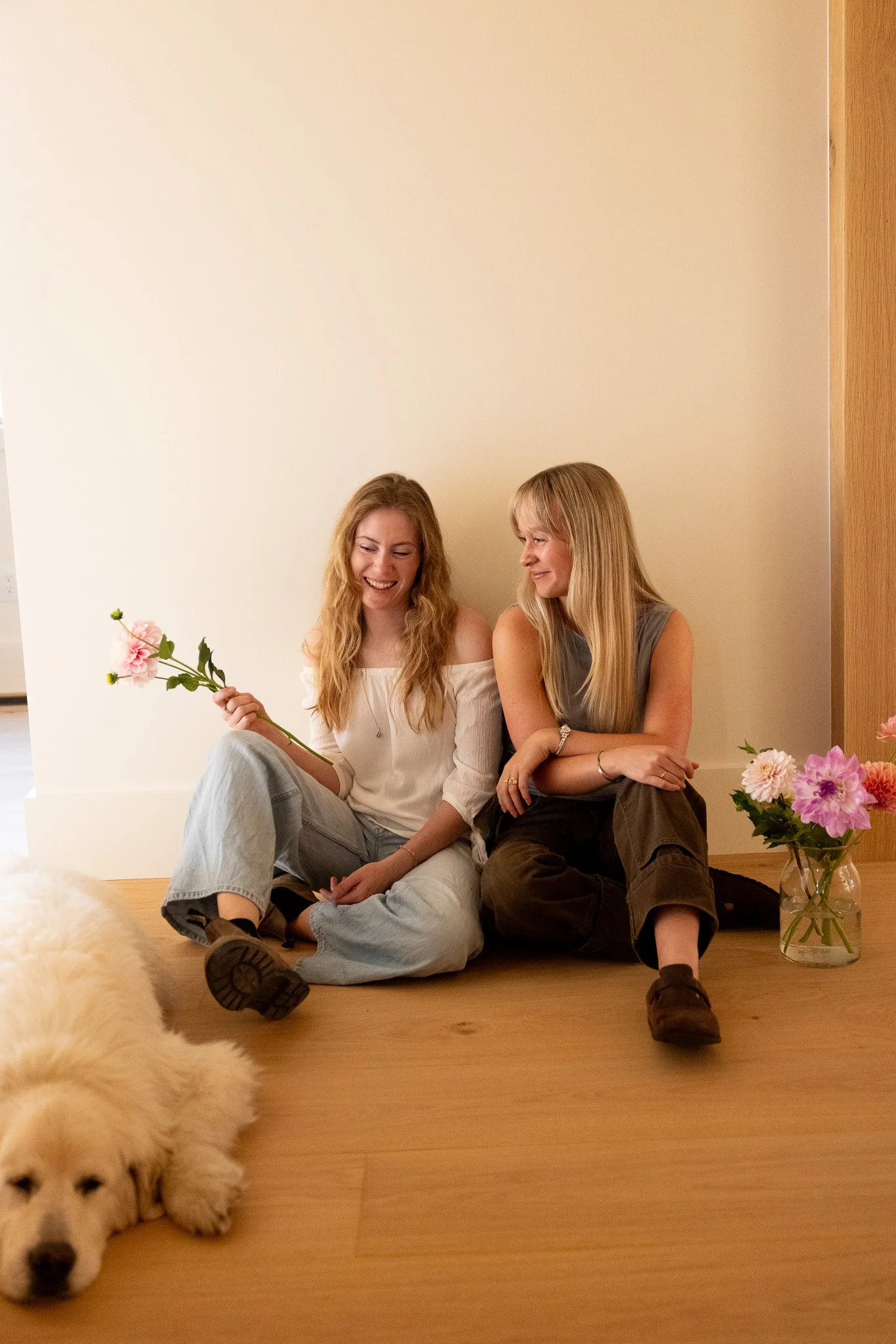 Two women sitting on the floor, smiling and engaging with each other, with a dog lying in the foreground. There are pink flowers in a vase behind them and a plain wall in the background.