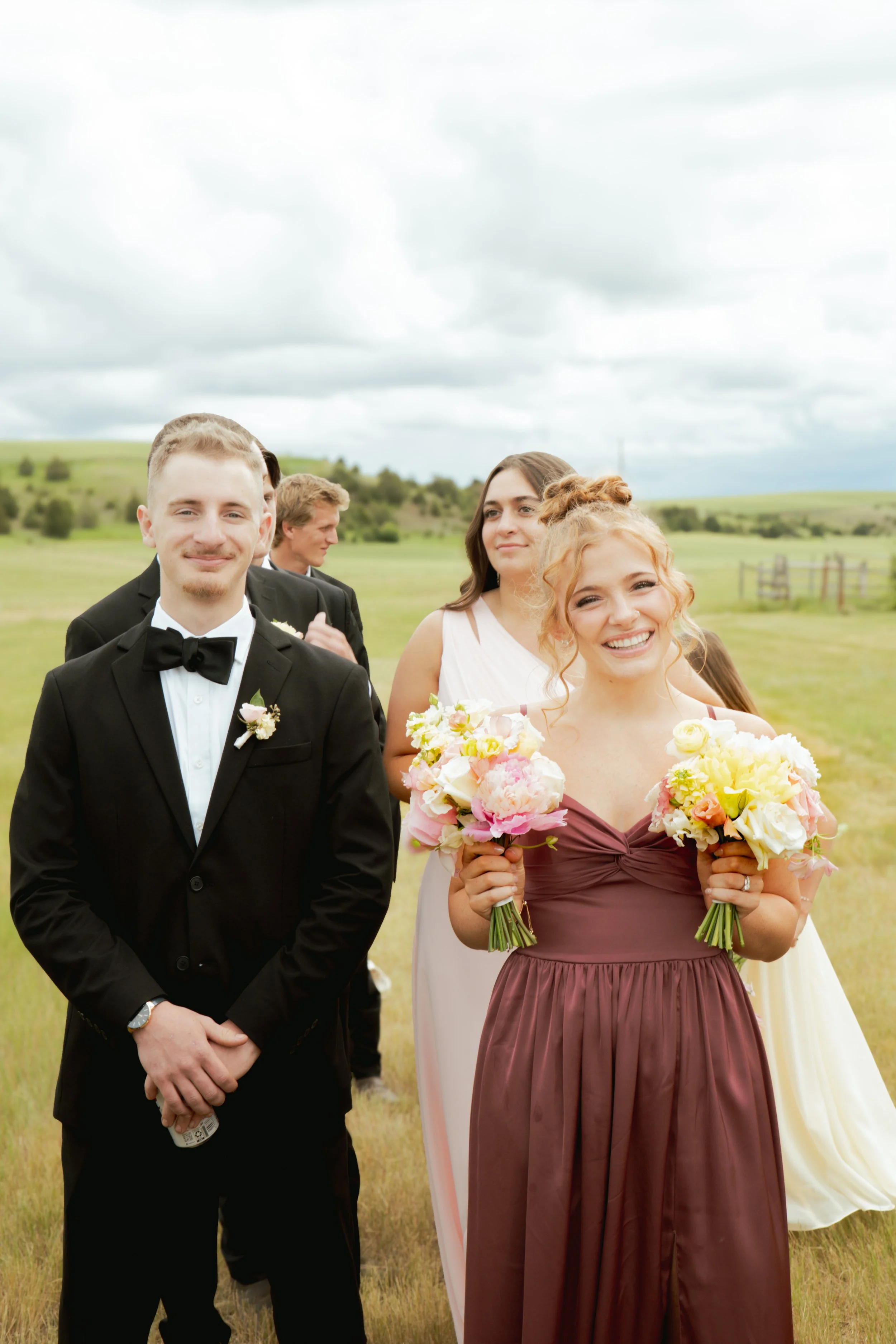 A wedding party outdoors on a grassy field with cloudy sky in the background. There are multiple people dressed in formal attire, including a man in a black tuxedo with a bow tie, a woman in a convertible dress holding a bouquet of flowers, and other