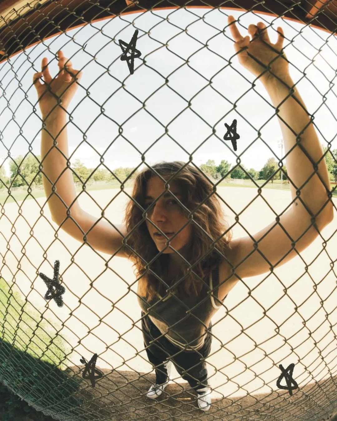 A young woman with curly hair standing behind a chain-link fence outdoors, gripping the fence with both hands, looking through it.