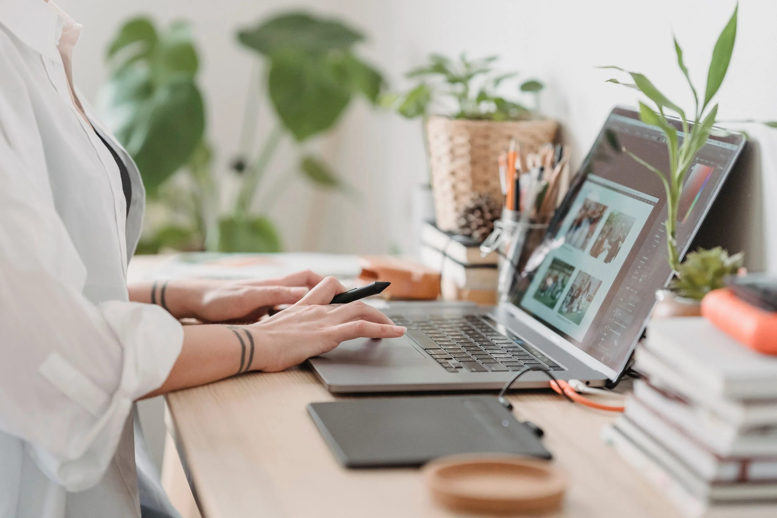 Person working on a laptop at a desk with plants and office supplies, using a stylus or pen.