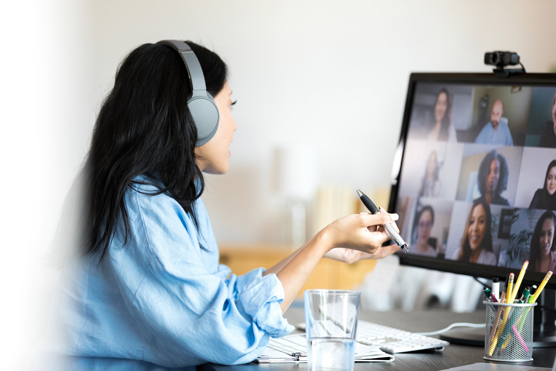 Woman with long dark hair wearing gray headphones working on a video call at a desk, with multiple group photos on the computer screen.