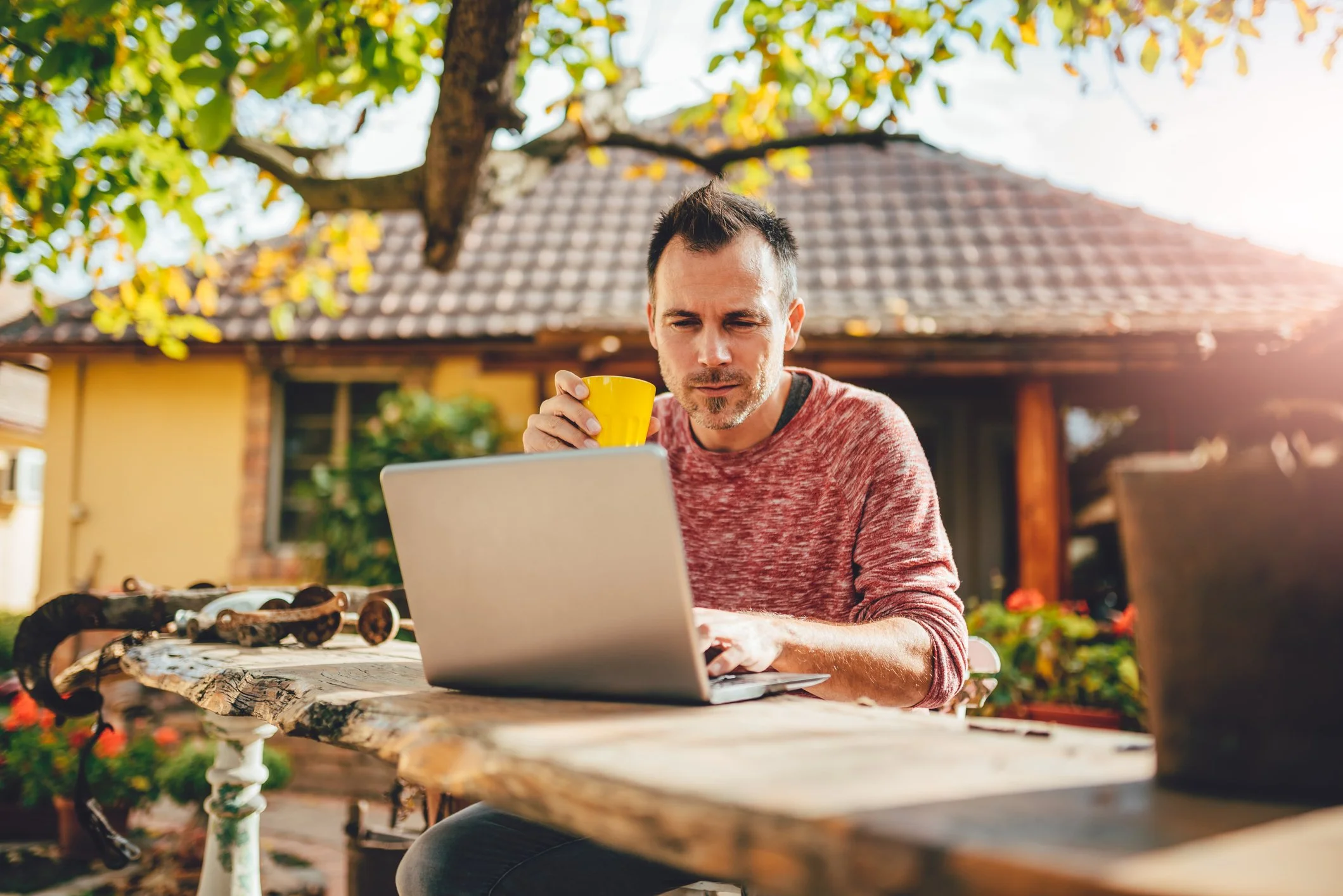 A man sitting at a rustic outdoor table holding a yellow mug and looking at a laptop with sunglasses placed on the table. There is a house and trees in the background on a sunny day.