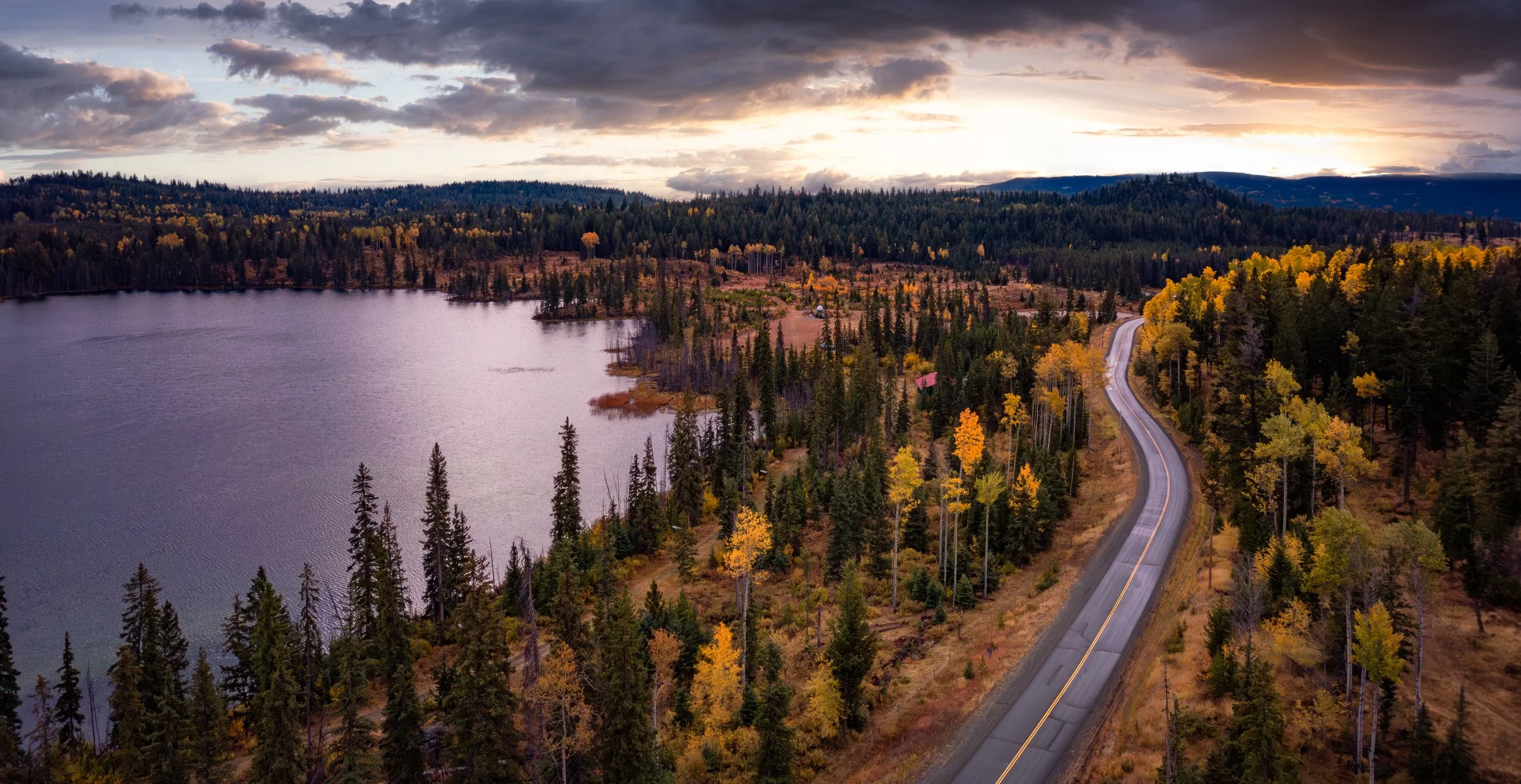 A scenic view of a lake surrounded by a forest with autumn-colored trees, with a winding road running alongside.