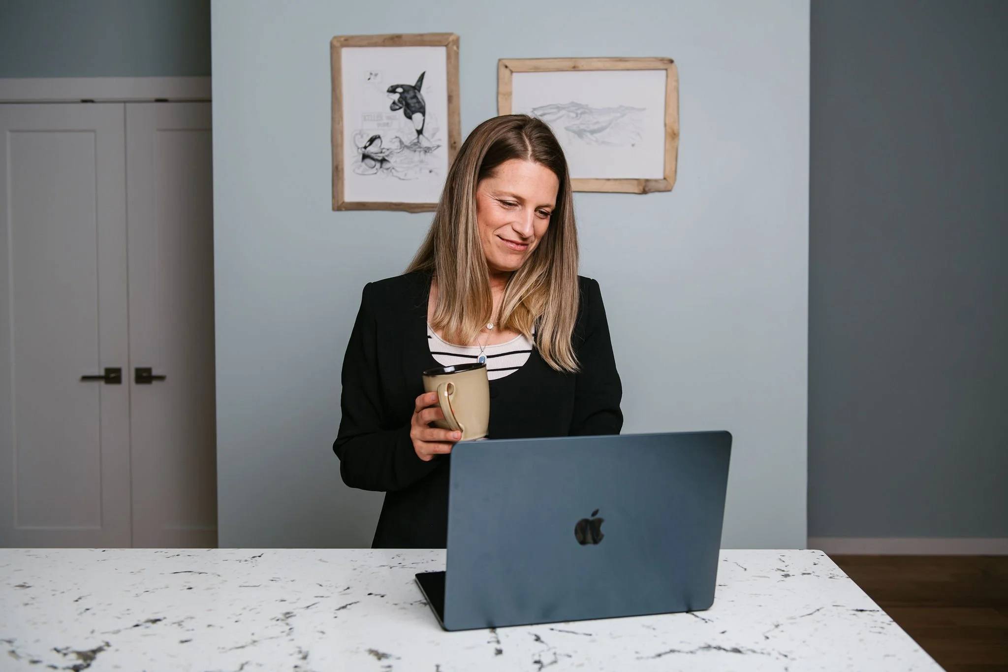 Woman in black blazer and striped shirt smiling and looking at her phone while holding a mug, standing behind a laptop on a white marble kitchen island.
